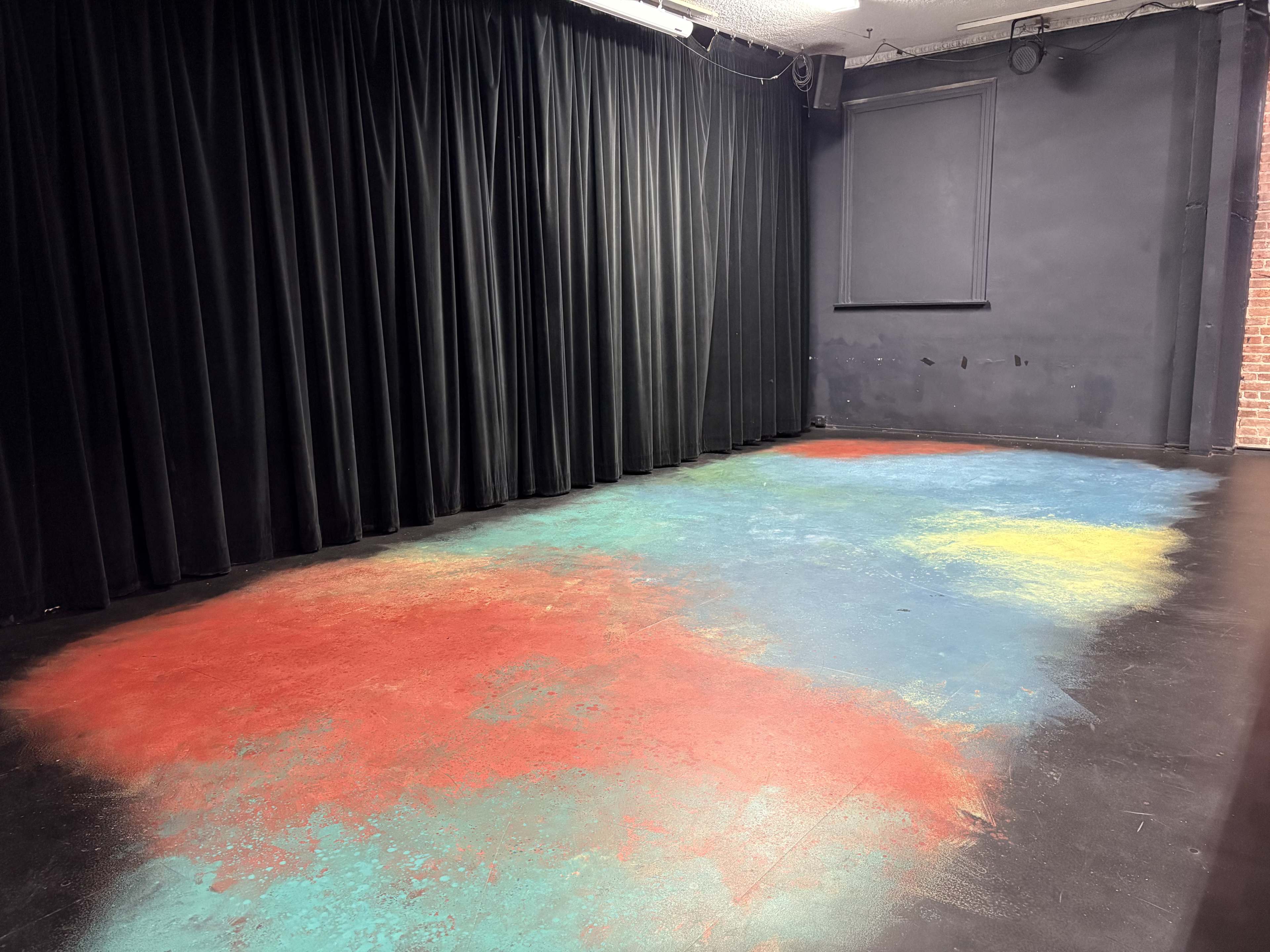 The image shows a performance space with a black curtain on one side and a floor painted in a splattered pattern of red, blue, and yellow.