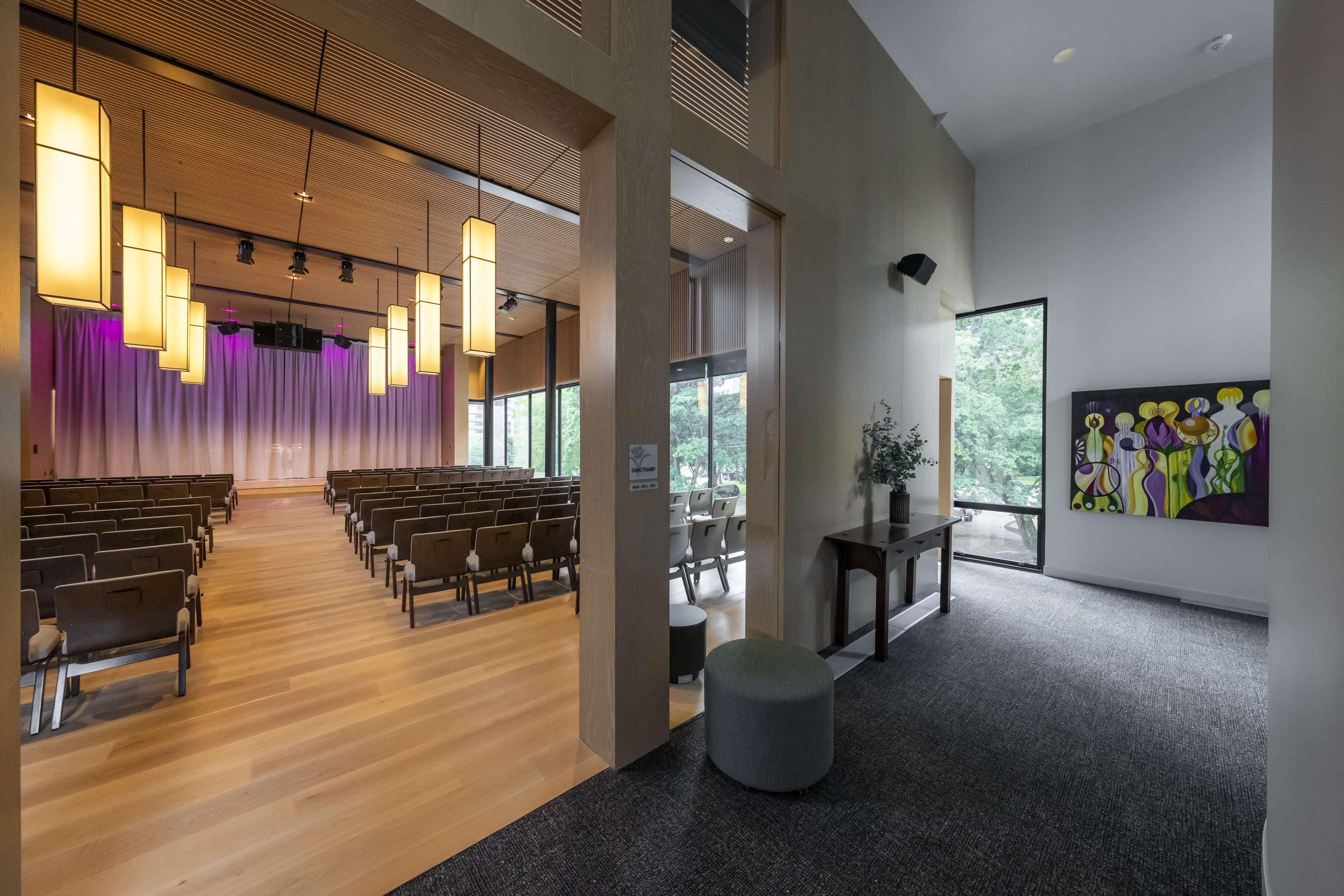 A well-lit entryway leads to a spacious auditorium with rows of seats facing a stage, adorned with pendant lights and purple curtains.