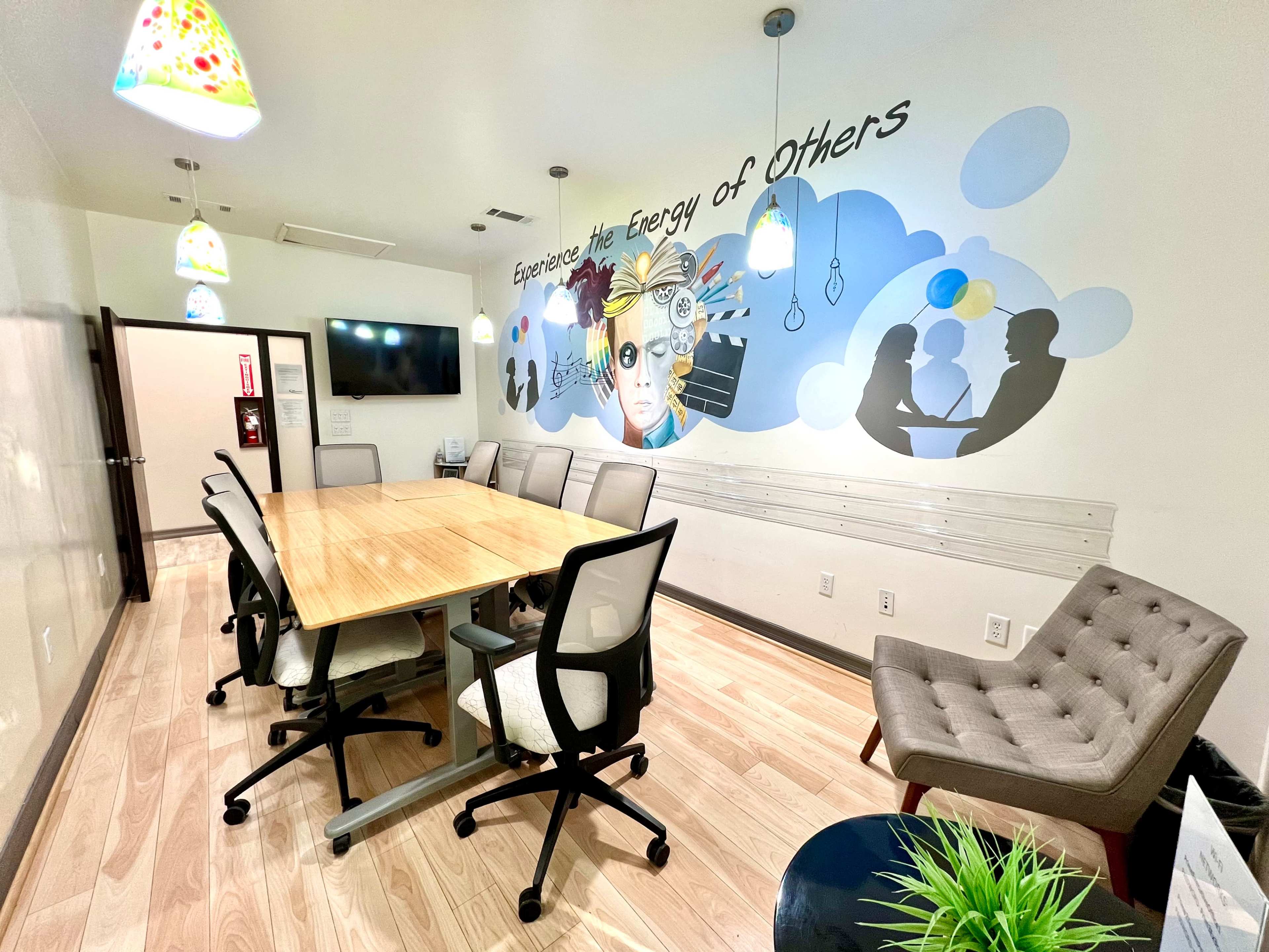 The image shows a modern conference room with a large wooden table surrounded by chairs, featuring a mural on the wall that promotes collaboration and creativity.