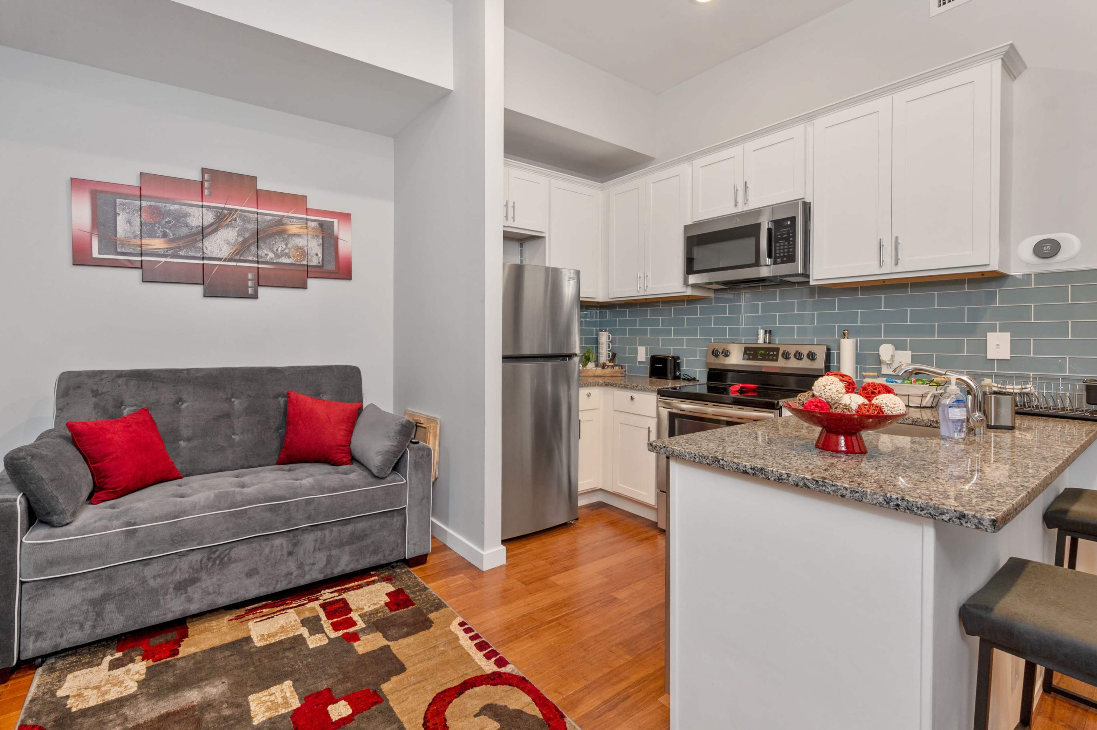 A modern kitchen and living area feature light cabinetry, stainless steel appliances, and a gray sofa with red accents.