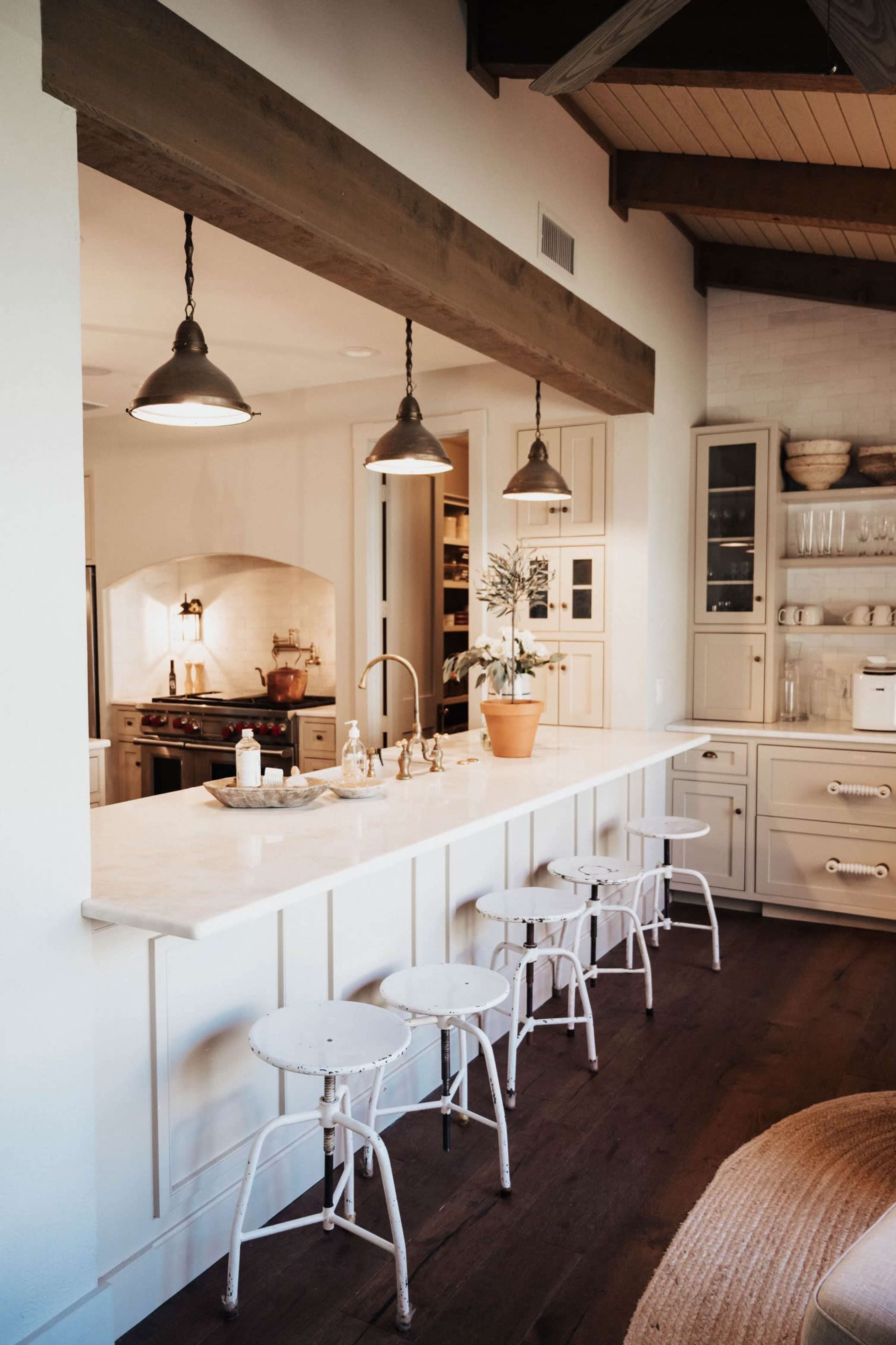 A modern kitchen features a long white countertop with three metal stools and pendant lighting above.