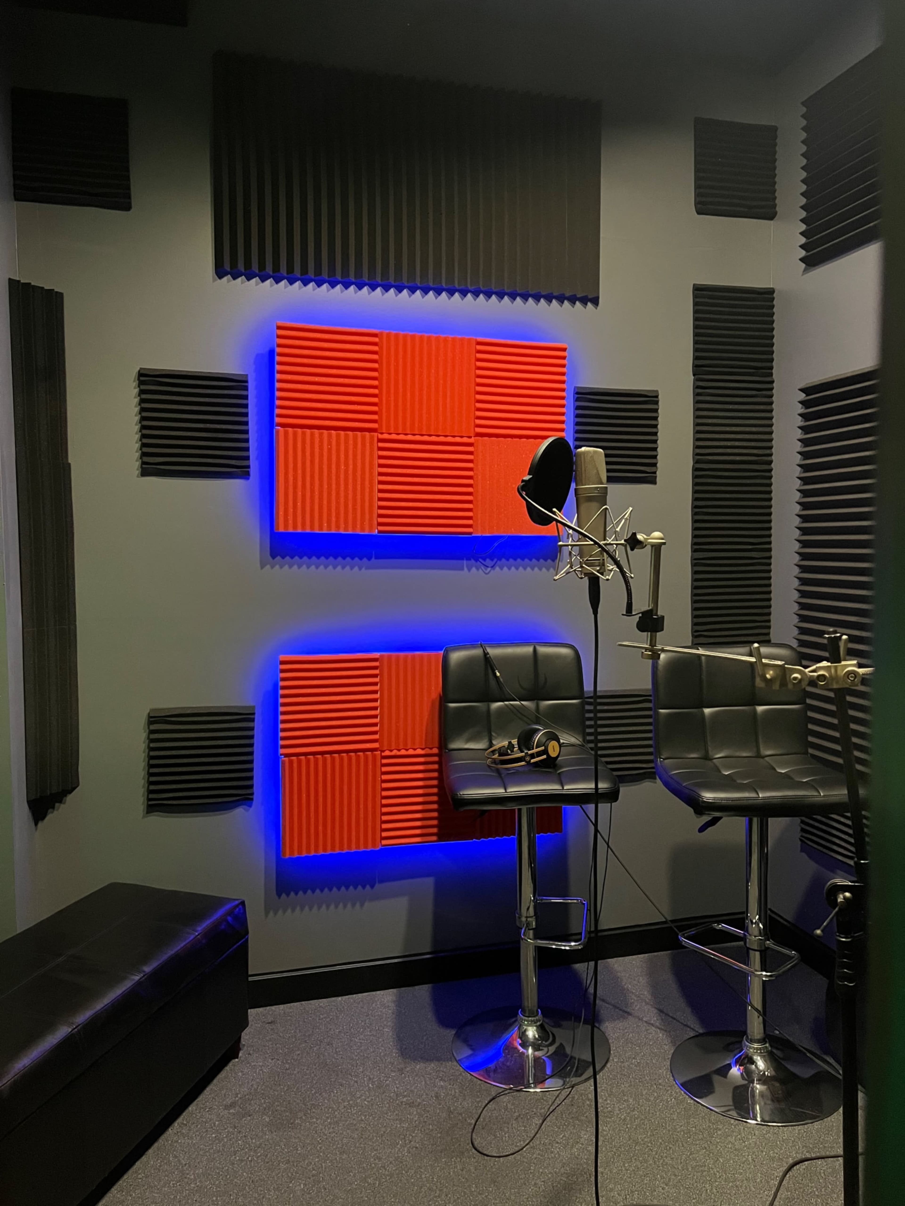 A soundproof recording studio features black and red acoustic foam panels, a microphone on a stand, and two black stools.
