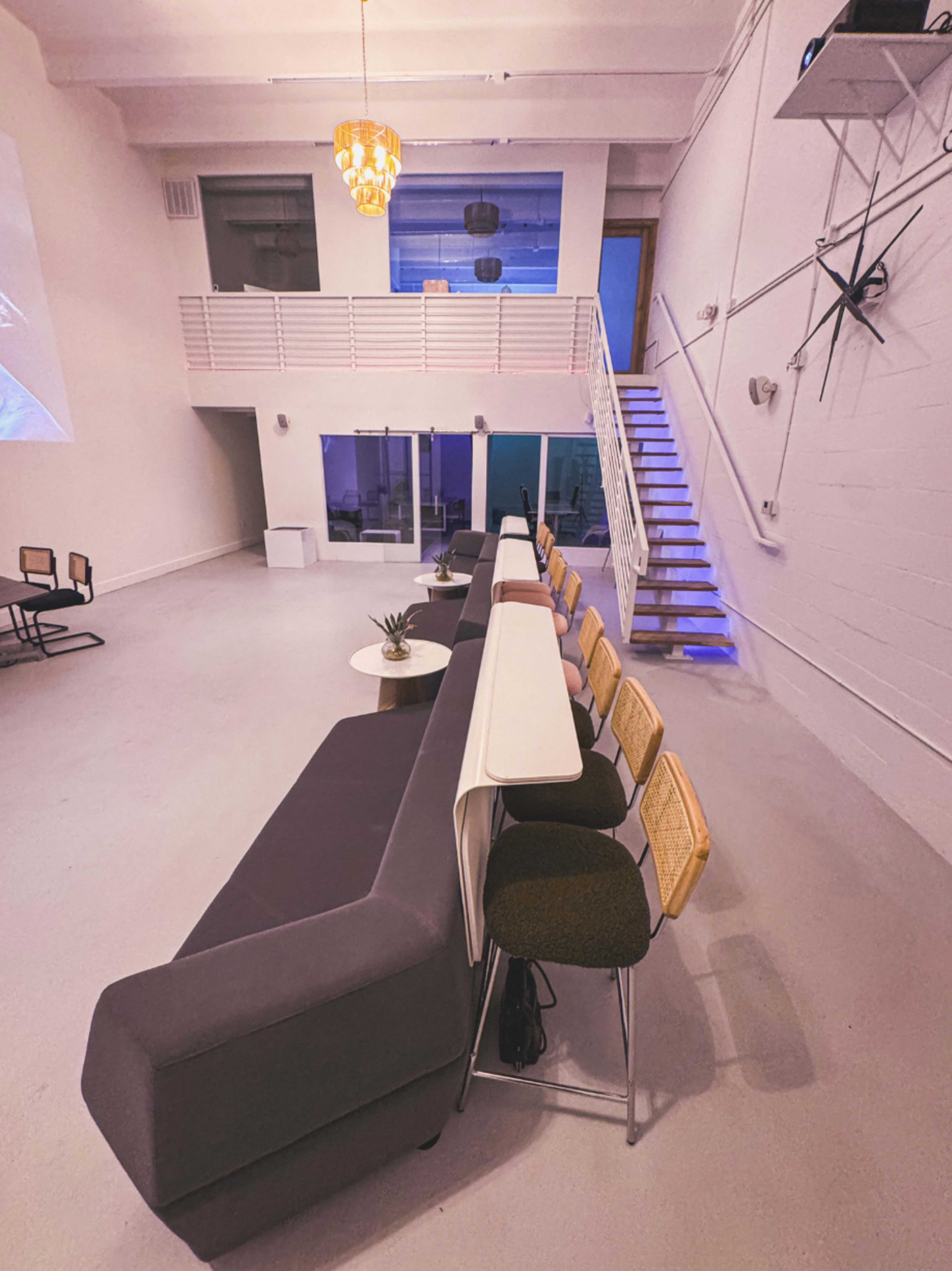 The image shows a modern, open interior space featuring a gray sectional sofa, bar stools, and a stairway leading to an upper level.
