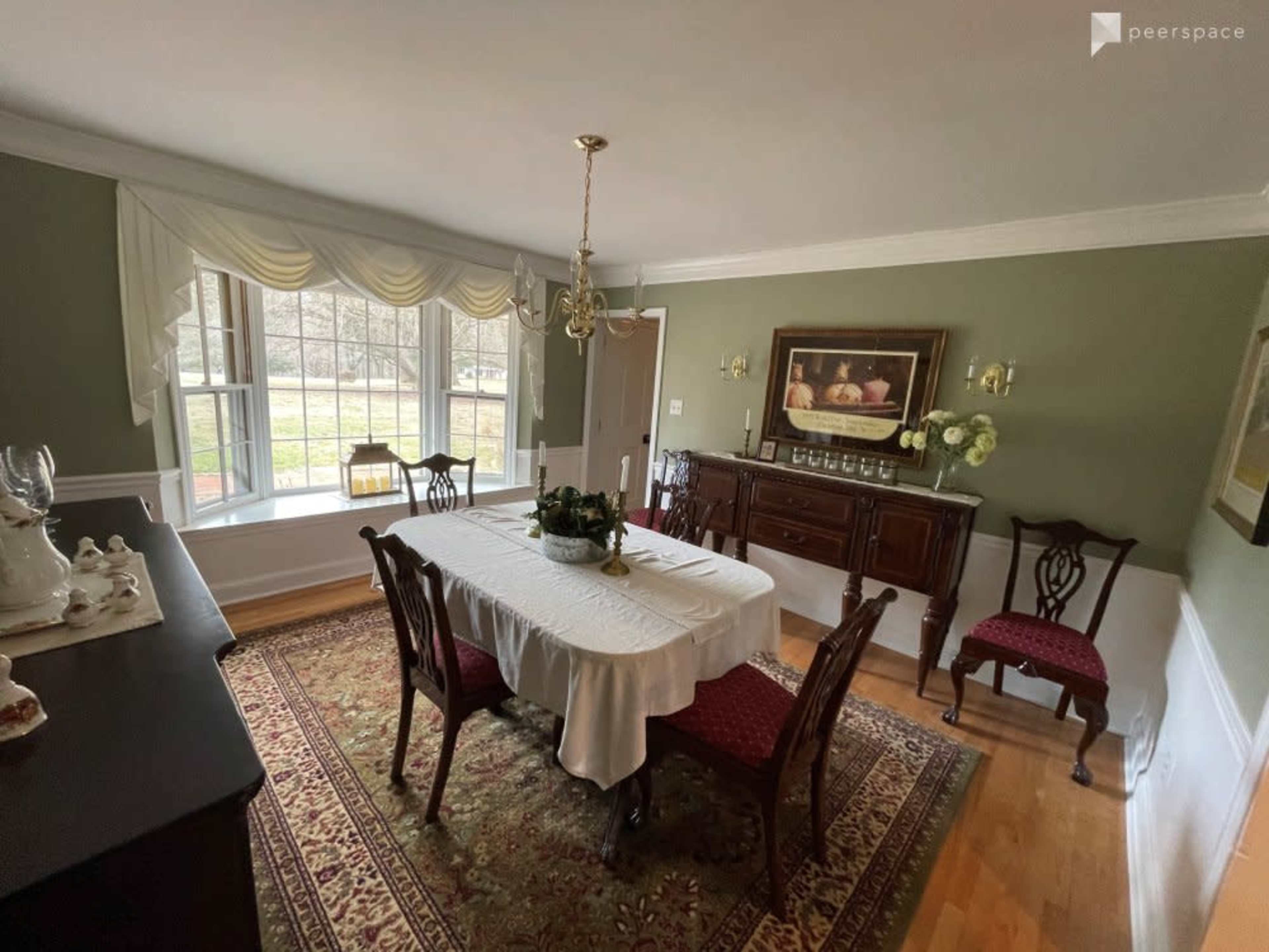 A dining room features a table with a tablecloth, surrounded by chairs, and a large window with curtains overlooking a green landscape.
