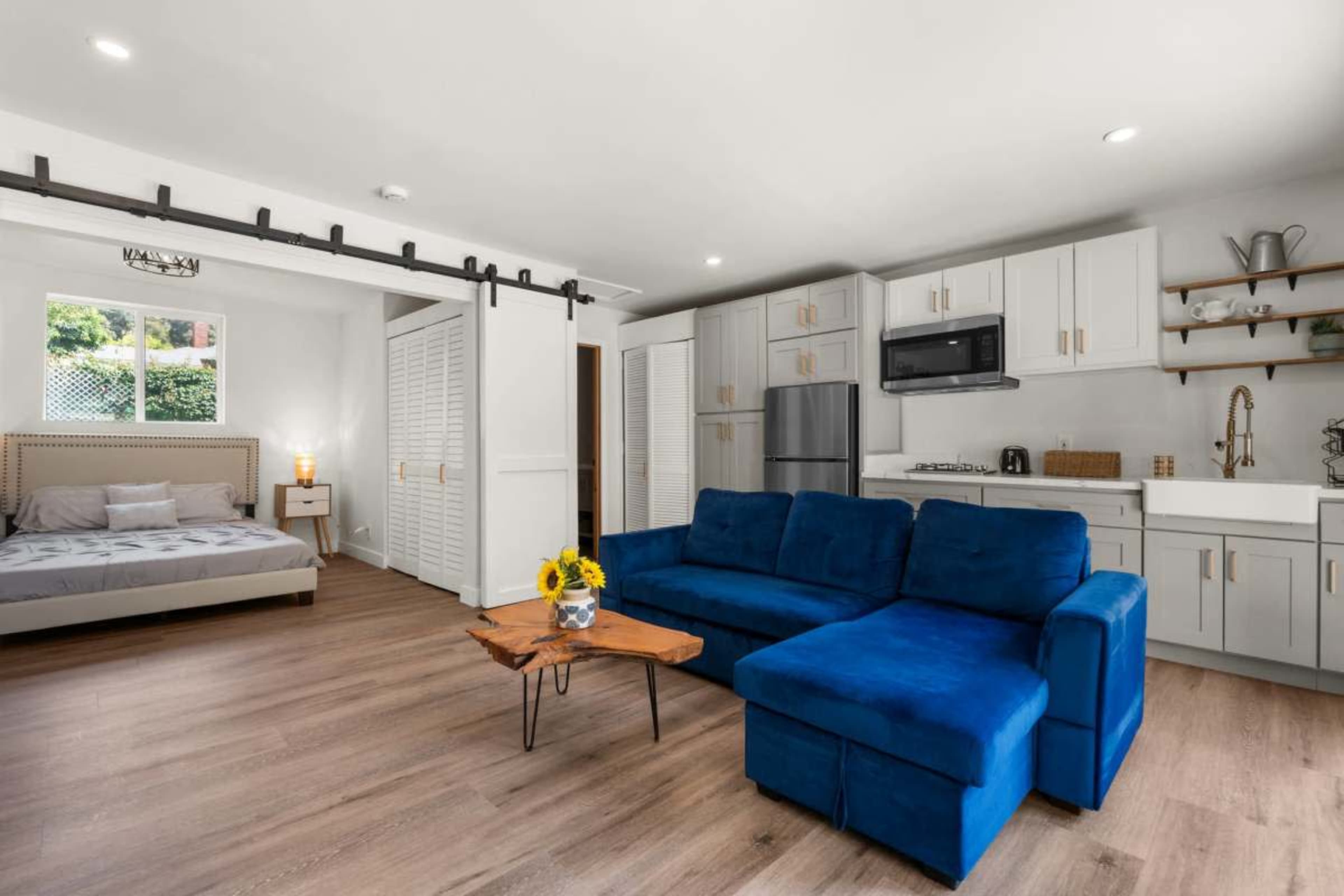 The image shows a modern studio apartment featuring a blue sofa, a wooden coffee table with sunflowers, a bed in the corner, and a kitchenette with white cabinets and stainless steel appliances.