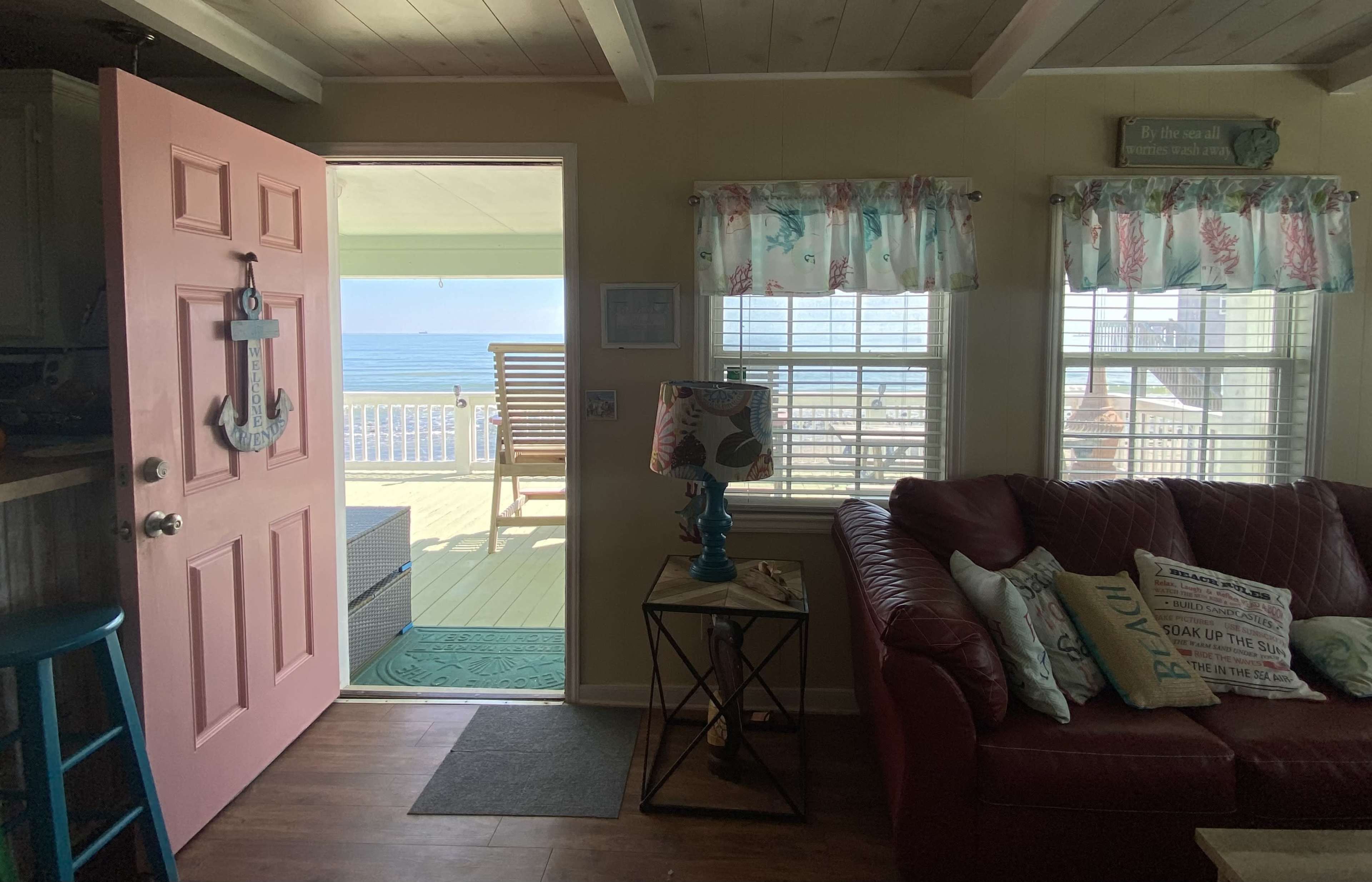 The image shows a living space with a pink front door leading to a deck overlooking the ocean, featuring a red couch and light-colored curtains.