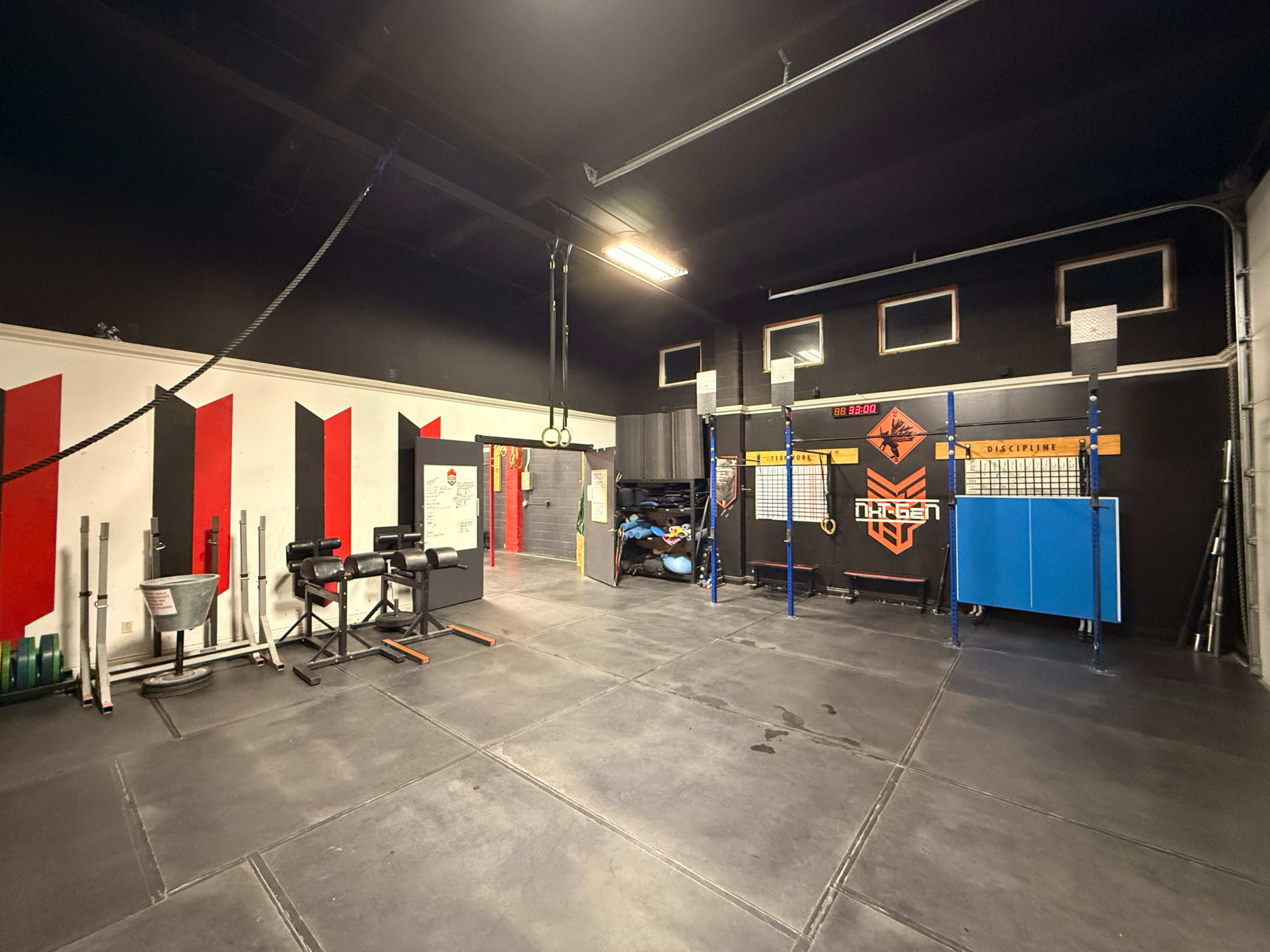The image shows an interior of a gym featuring various exercise equipment, including weights and exercise mats, in an organized space with a black and red color scheme.