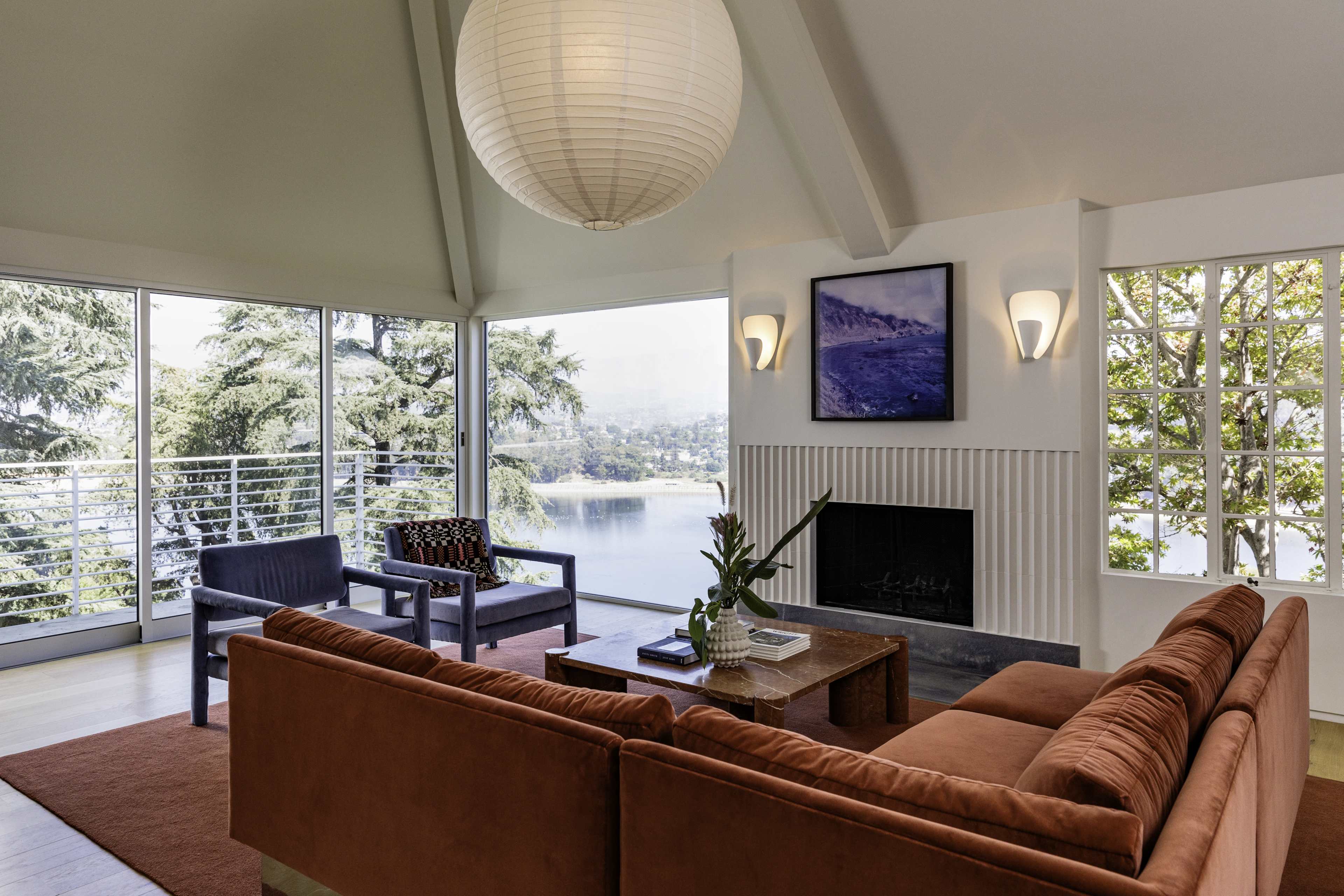A modern living room features a large, orange sofa, two chairs, and a coffee table, all with a view of a body of water through expansive windows.