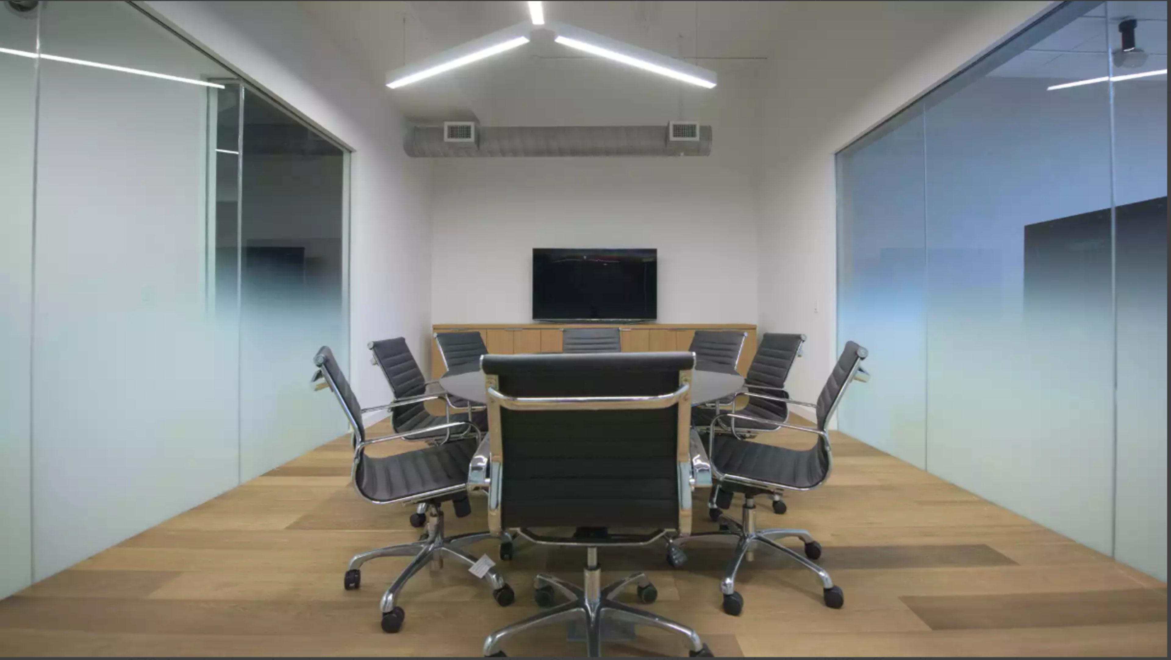 A modern conference room features a round table surrounded by adjustable chairs and a wall-mounted screen.