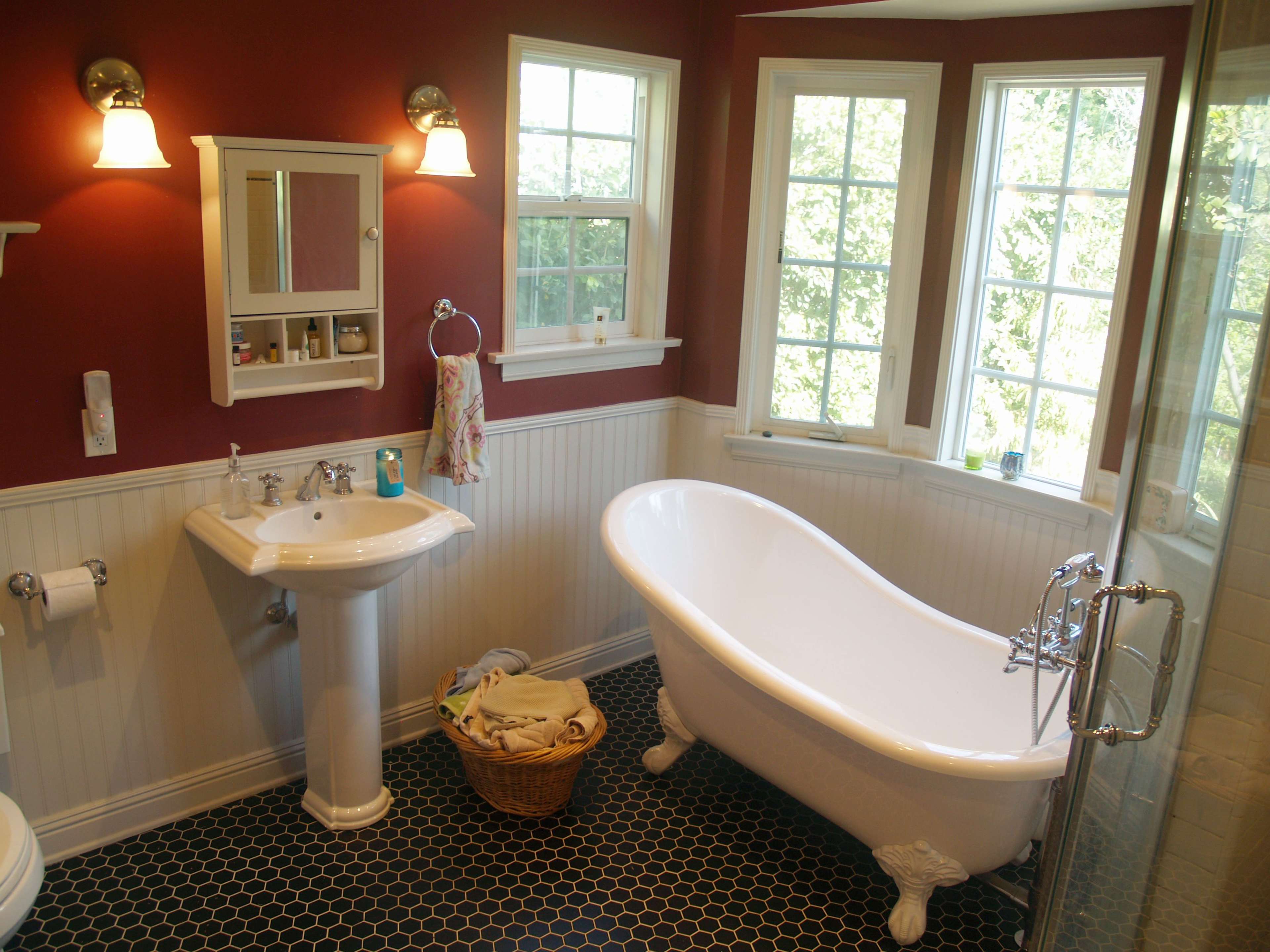 A bathroom featuring a freestanding white bathtub, a pedestal sink, and large windows allowing natural light to enter, with a dark hexagonal tile floor.