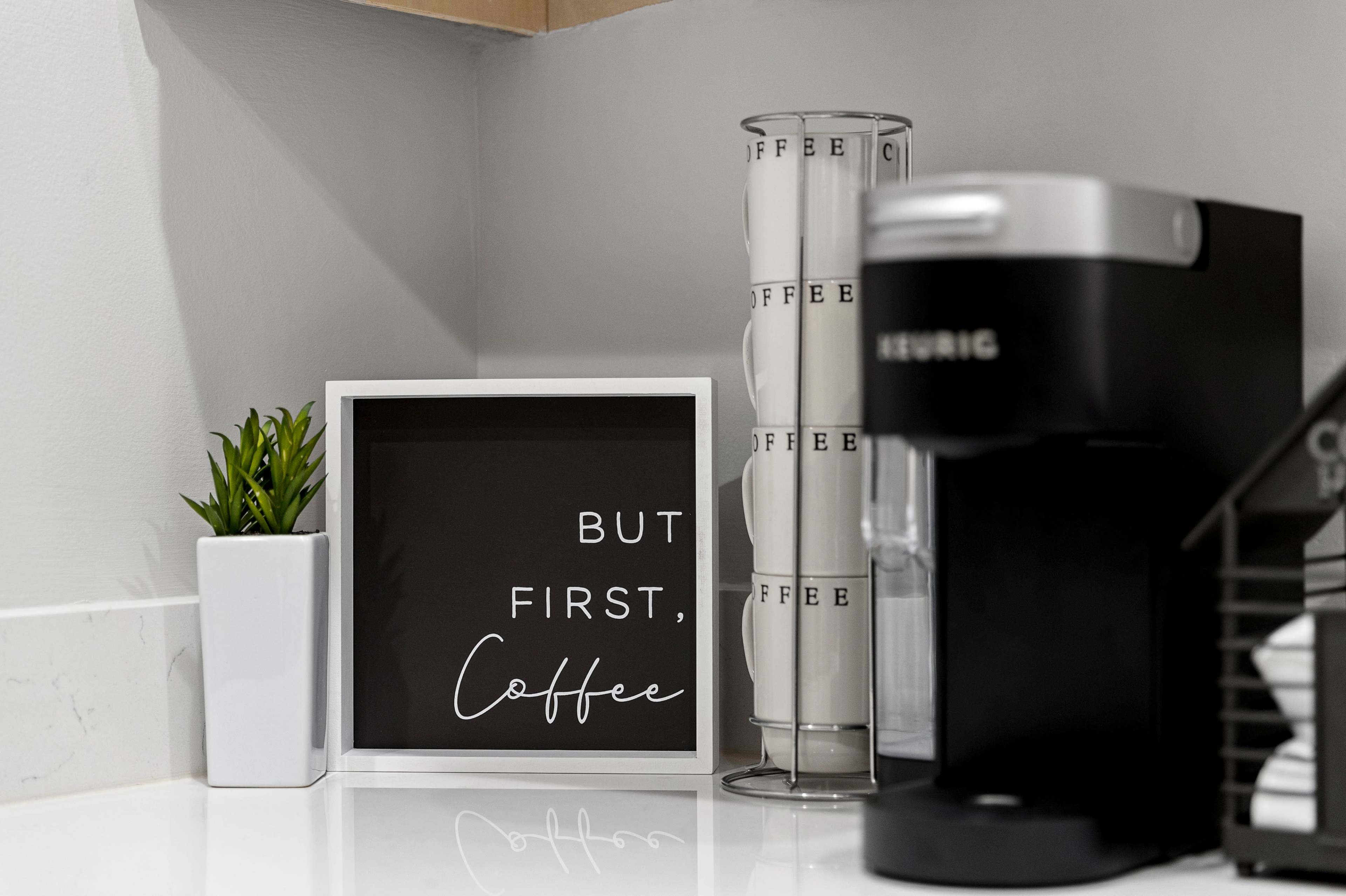 A coffee station features a black coffee maker, a decorative framed sign that reads "BUT FIRST, COFFEE," and a cylindrical container labeled "COFFEE."