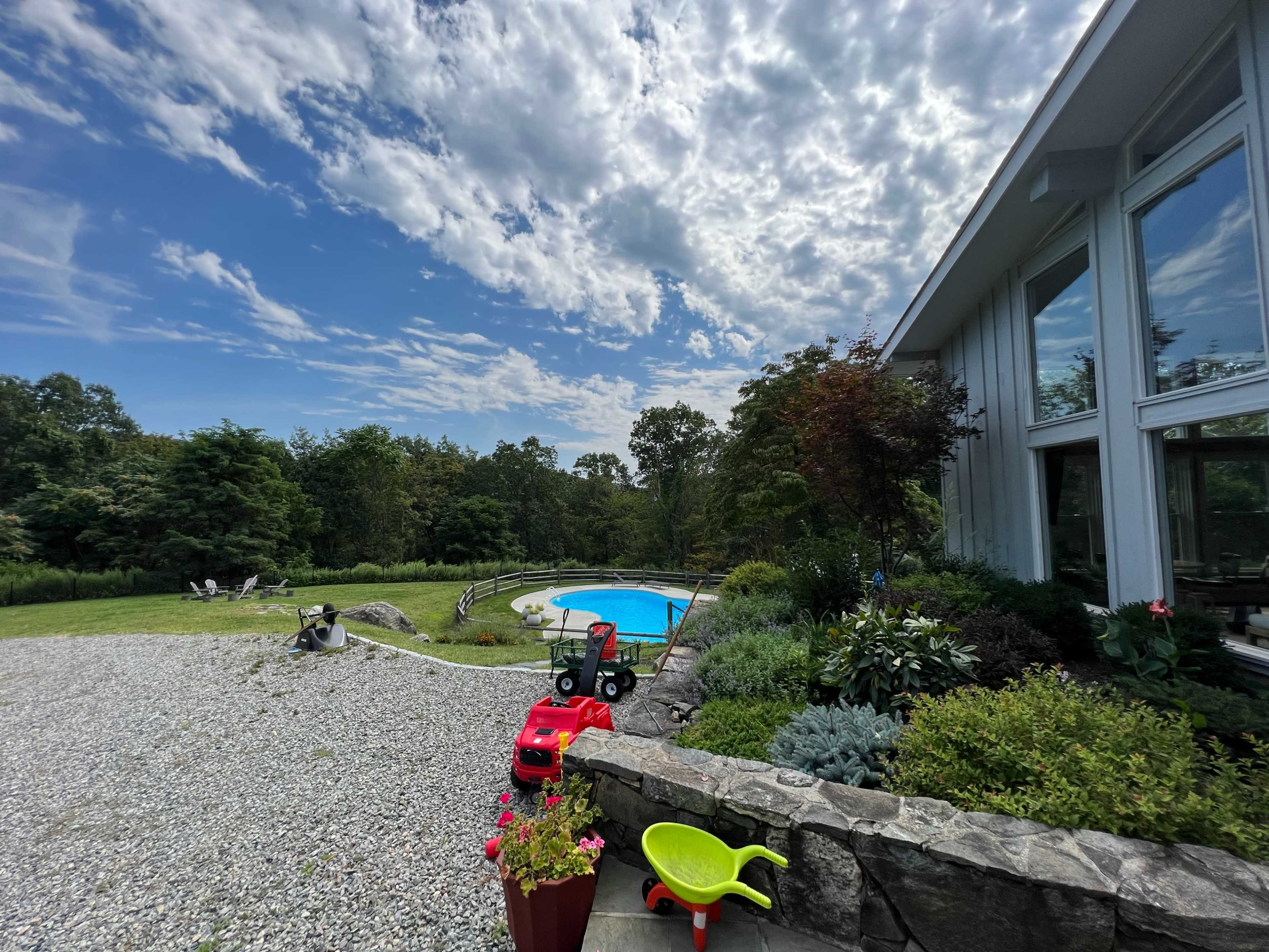 A landscaped backyard features a swimming pool surrounded by greenery, with a gravel area and a colorful toy truck parked nearby.