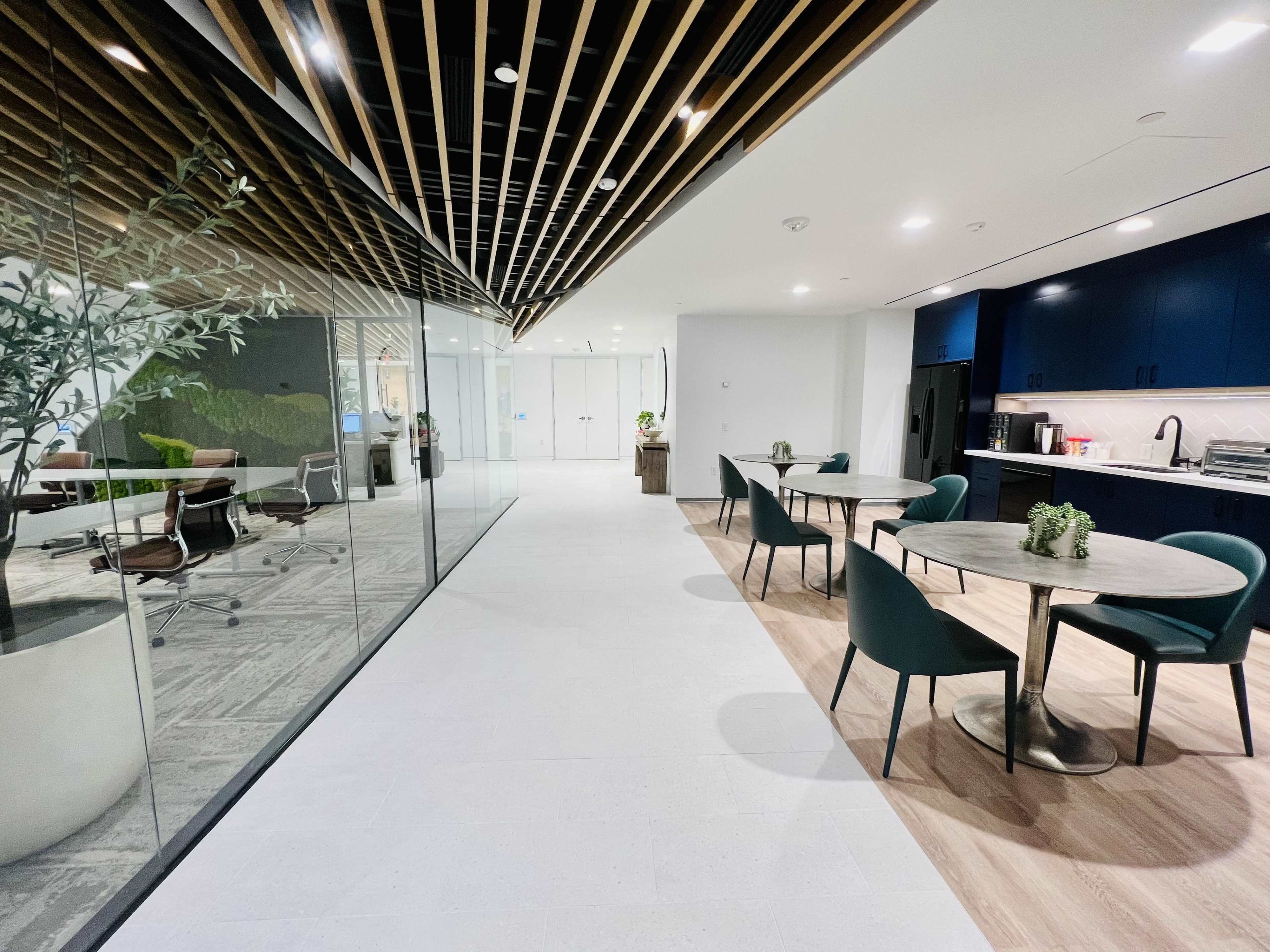 The image shows a modern office space featuring a kitchenette area, circular tables with green chairs, and a glass partition separating the work area.
