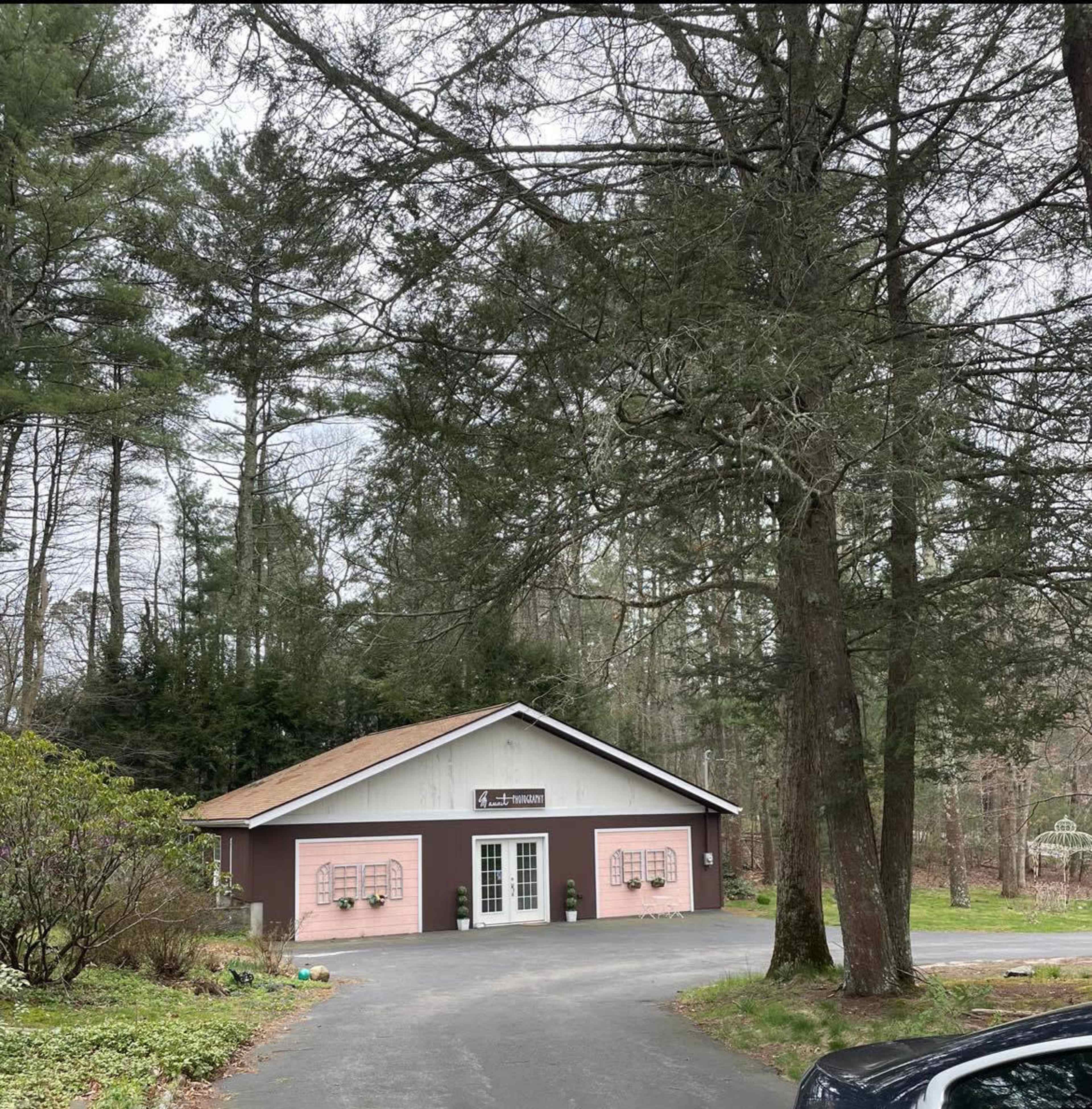 A two-toned building with pink lower walls and a white upper section is situated at the end of a driveway surrounded by tall trees and shrubs.