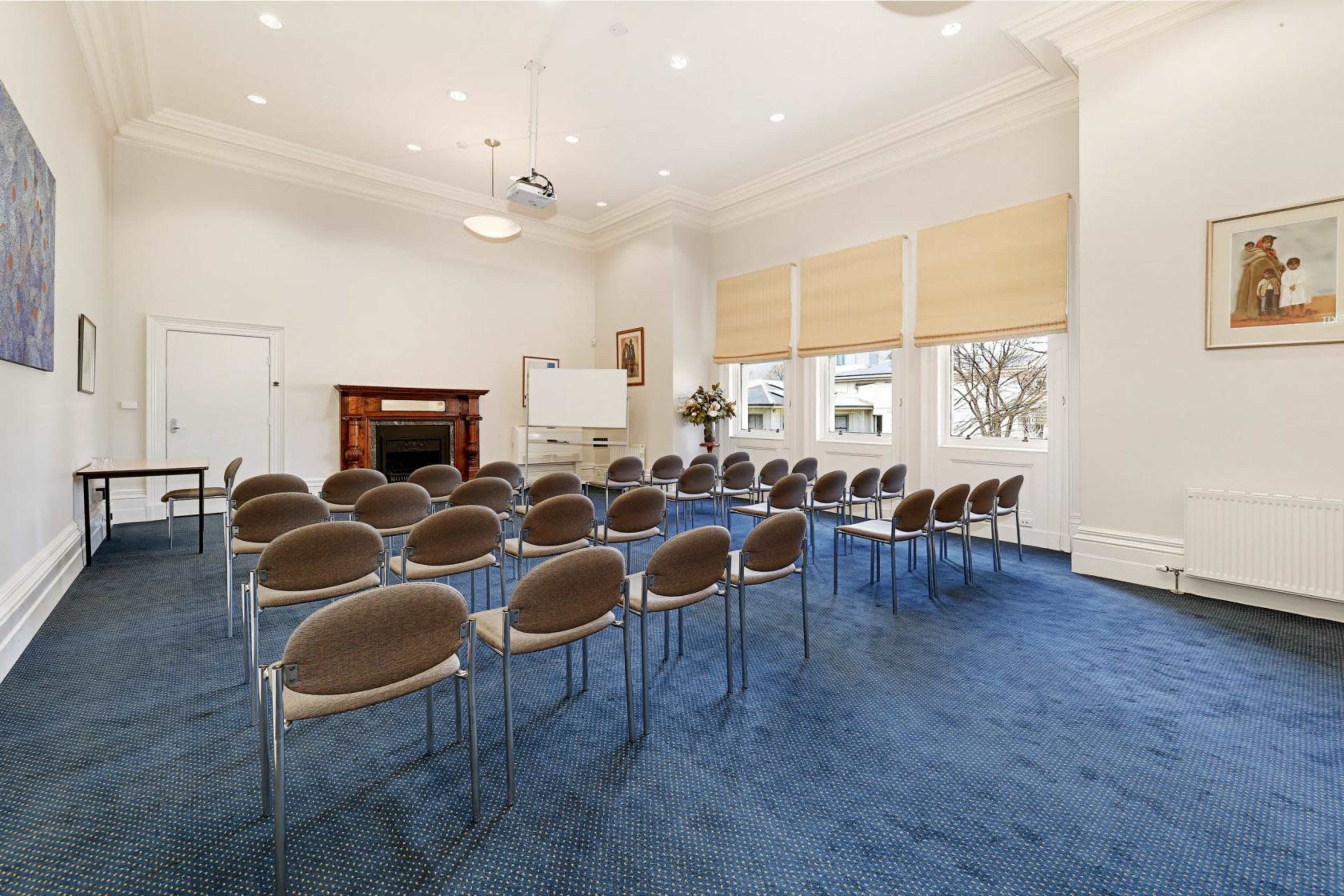 The image shows a meeting room with arranged chairs facing a presentation area, featuring a fireplace and large windows with shades.