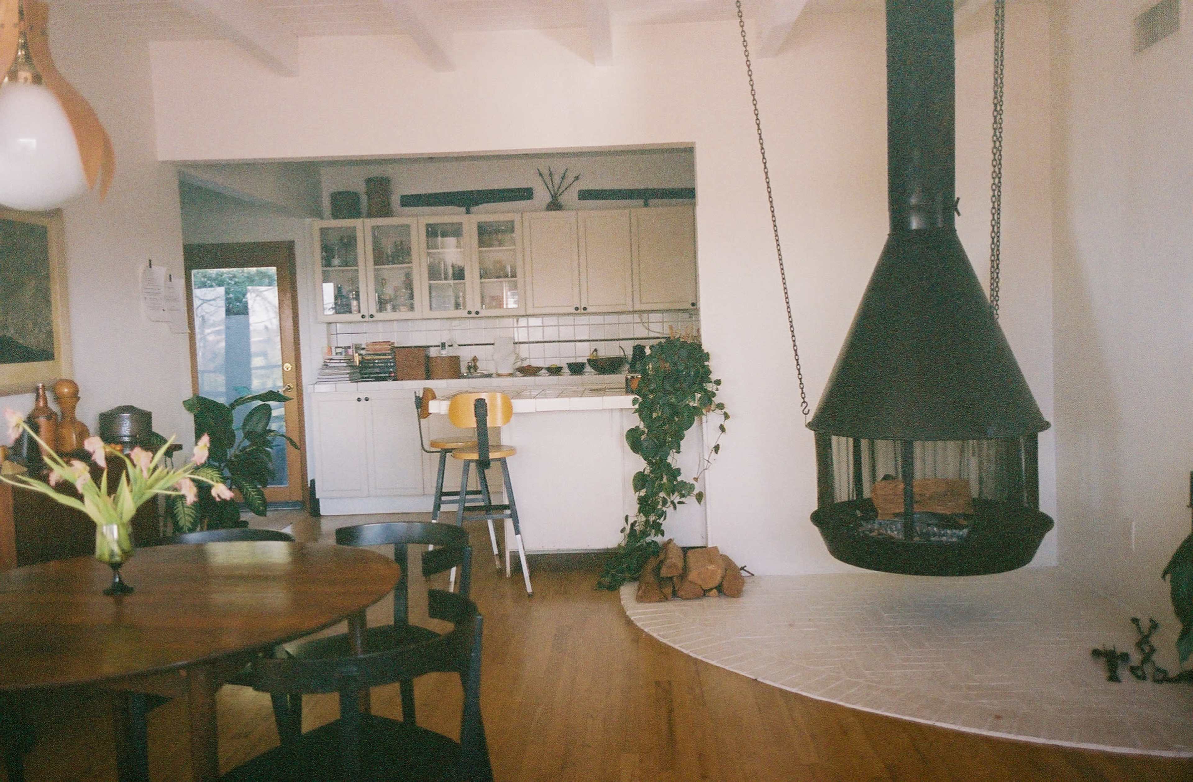 A cozy interior with a round wooden dining table in the foreground and a modern hanging fireplace nearby, while a kitchen area with white cabinets is visible in the background.
