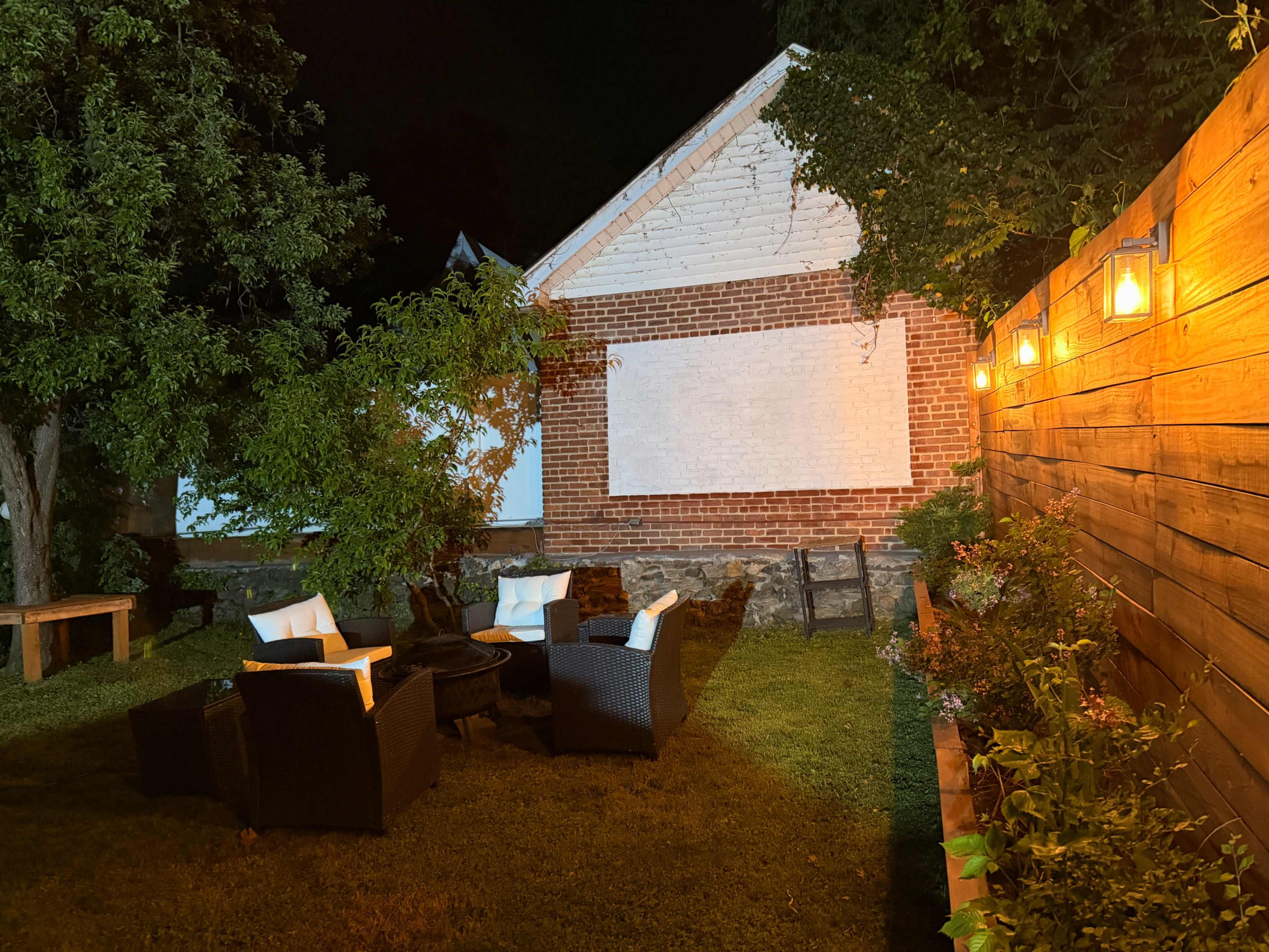 The image shows a dimly lit outdoor seating area with four wicker chairs and a table set on green grass, adjacent to a brick wall and lined with a wooden fence adorned with lights and plants.