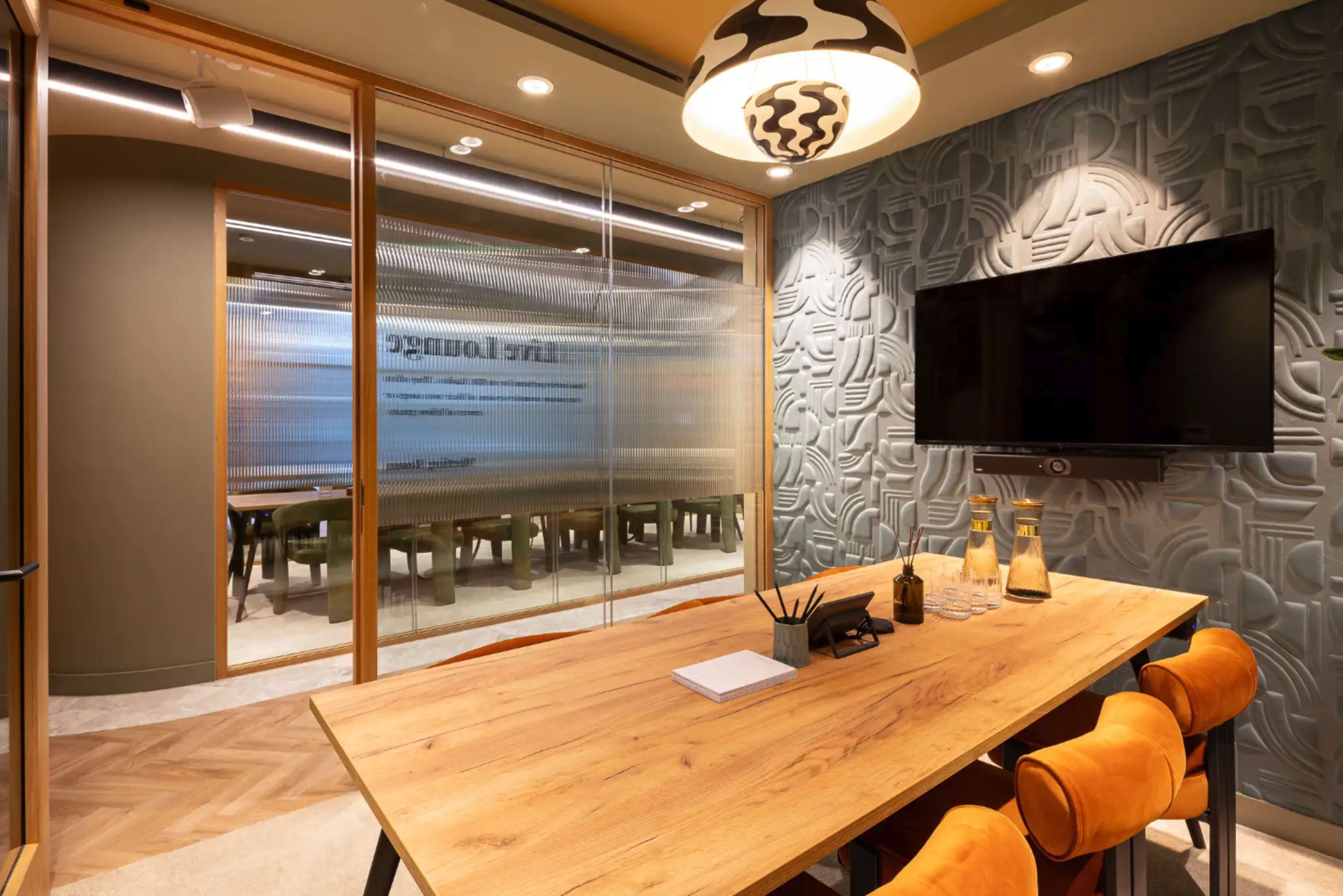 A modern conference room features a large wooden table surrounded by orange chairs, with glass walls and a mounted television.