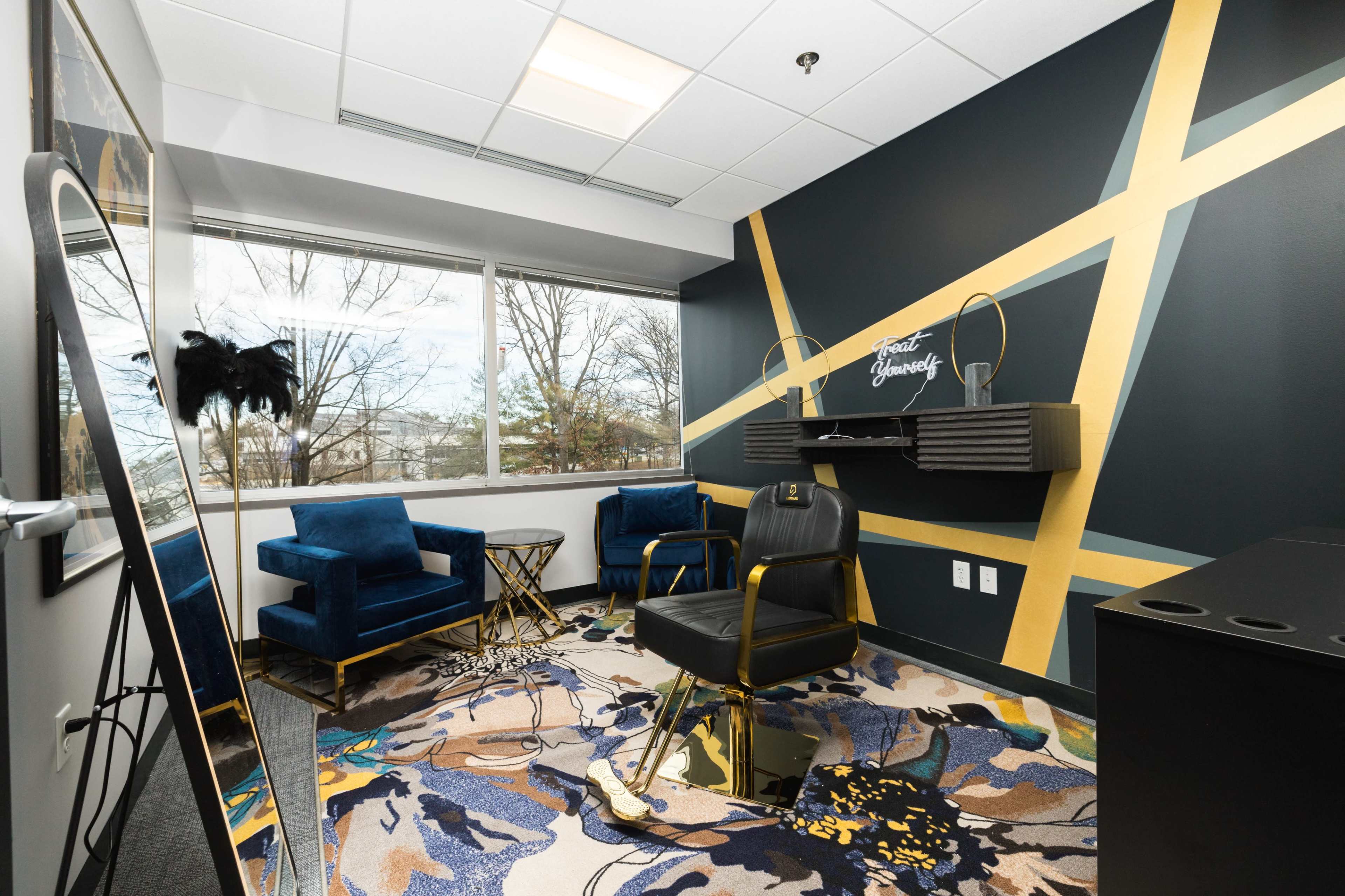 A modern office space featuring two blue armchairs, a black chair, and a decorative rug, with large windows allowing natural light and a minimalist black and gold color scheme on the walls.