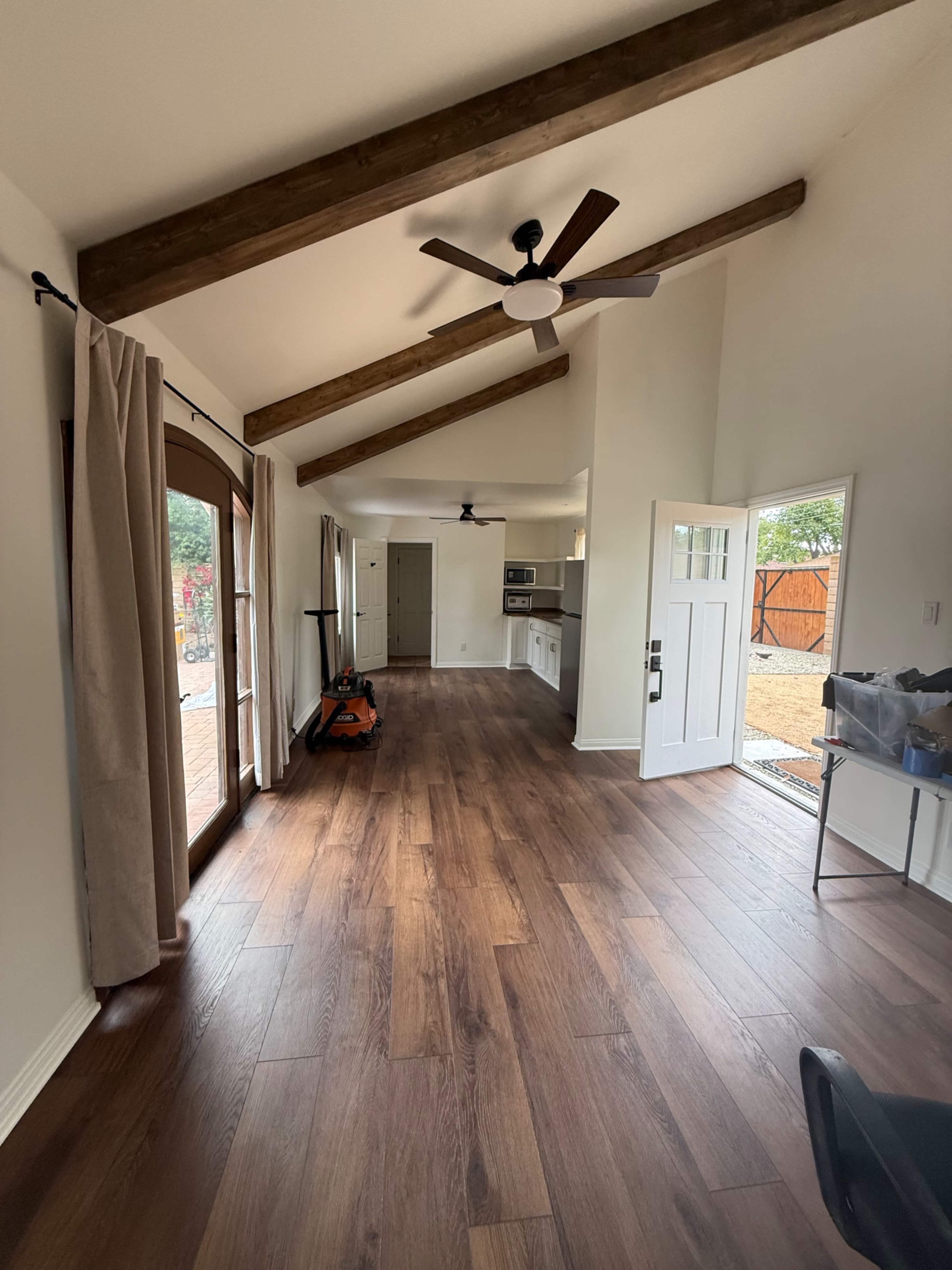 The image shows a spacious room with wooden beams, a ceiling fan, large windows, and wooden flooring, leading to a doorway that opens to an outdoor space.