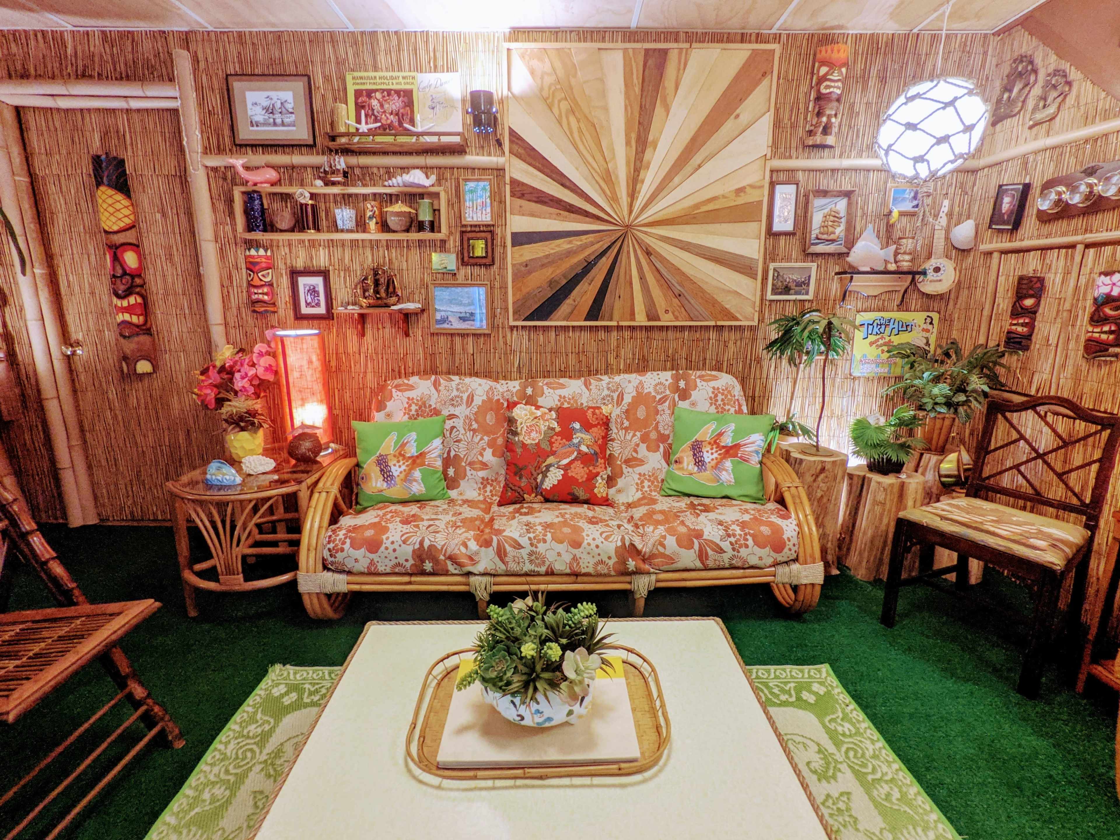 The image shows a tropical-themed living room with bamboo walls, a floral-patterned sofa, a wooden sunburst wall art piece, and various decorative items arranged on shelves.