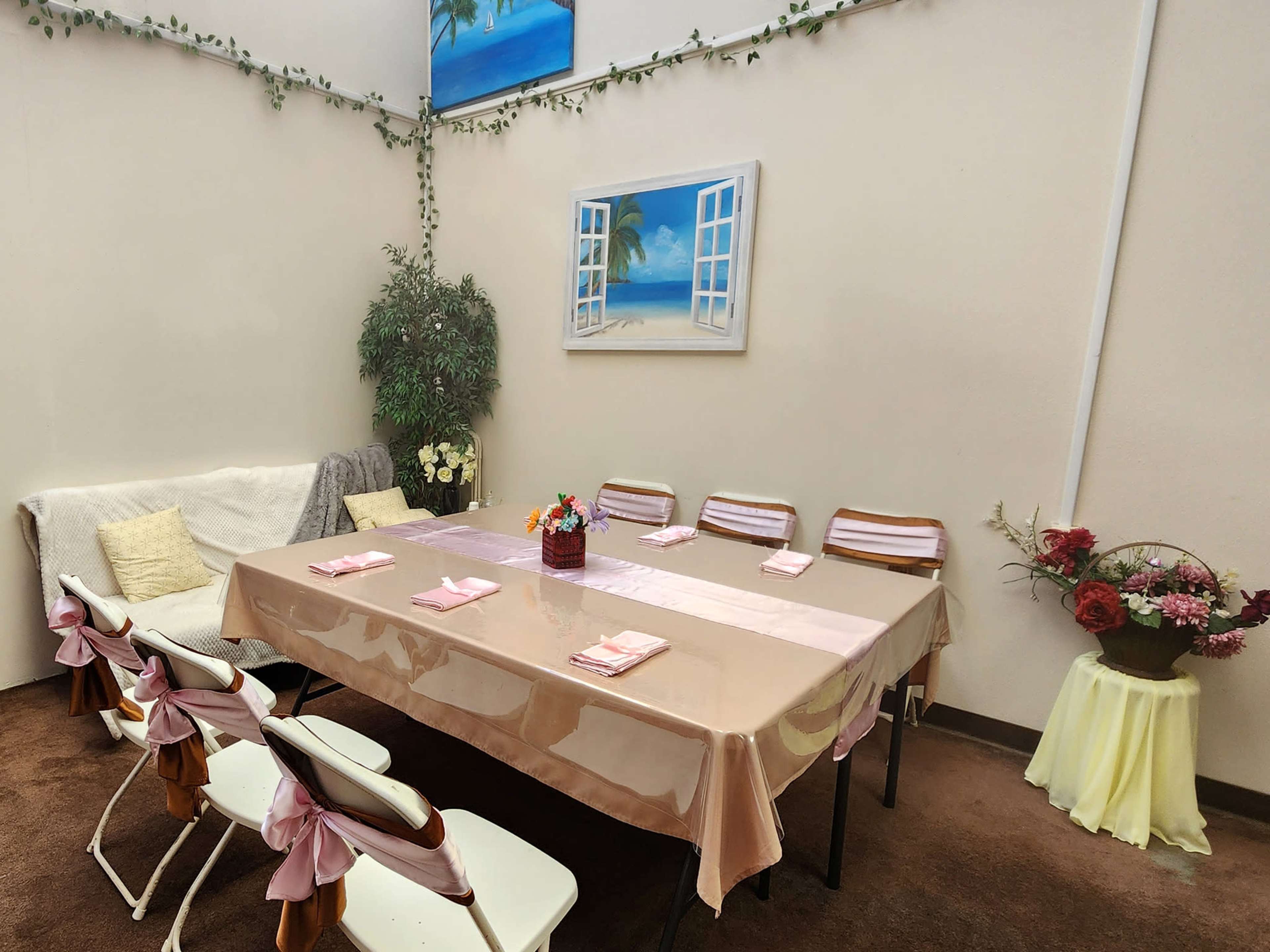A rectangular table is set with light pink tablecloth and napkins, surrounded by chairs, in a softly decorated indoor space with plants and wall art.