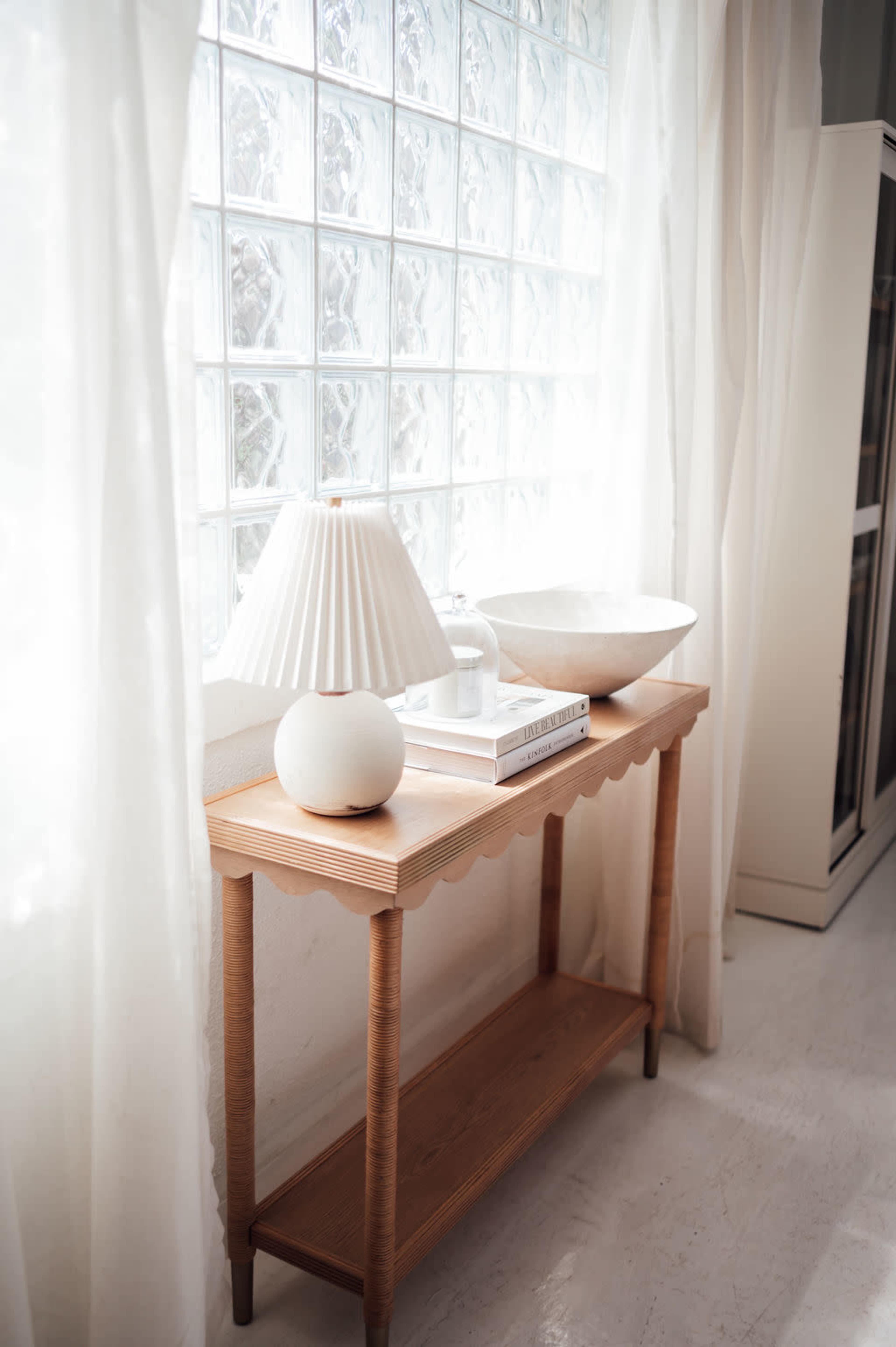 A wooden table with a lamp and a bowl sits beside a window with glass blocks, illuminated by natural light.