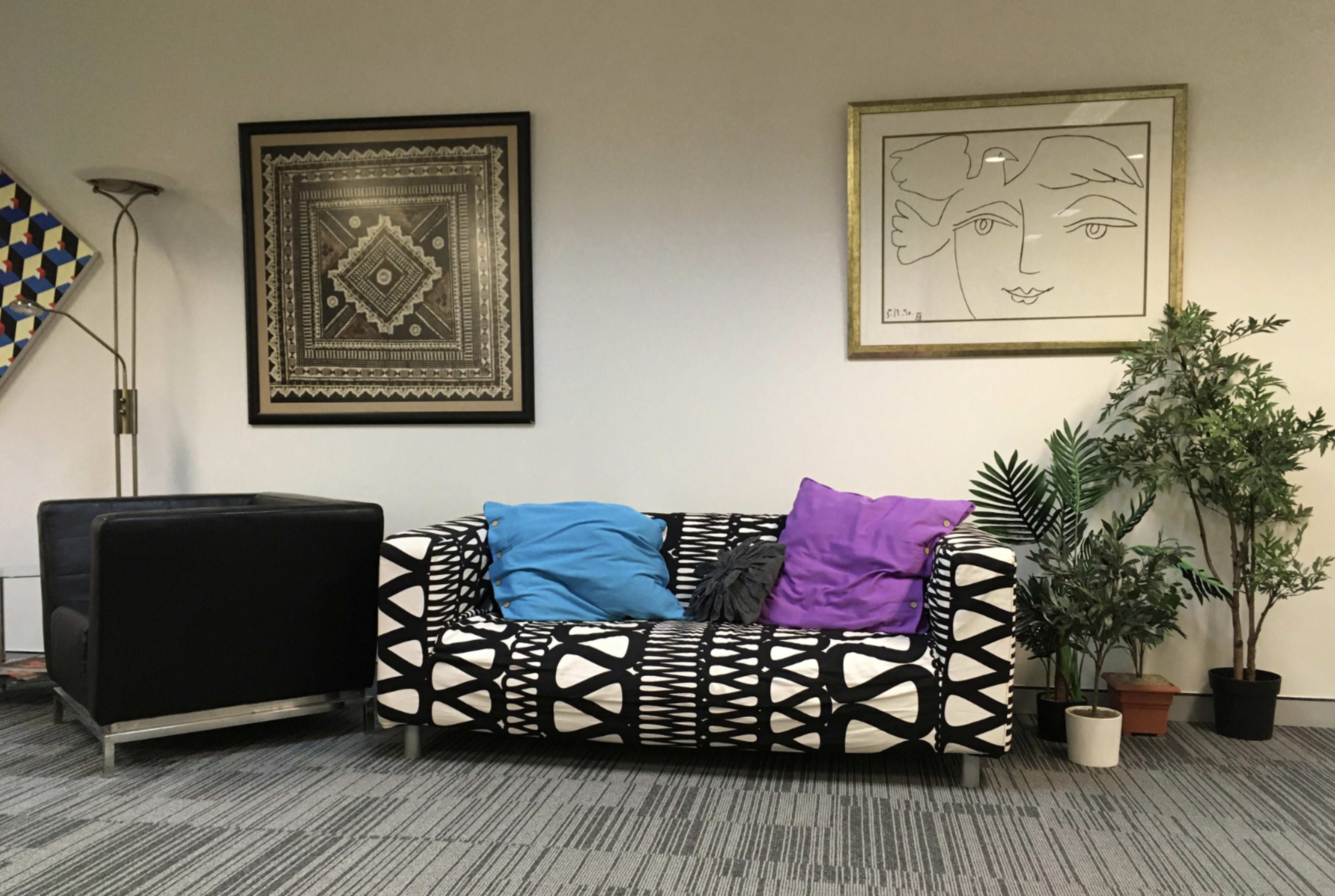 The image shows a living room corner with a patterned black and white couch adorned with blue and purple cushions, a black armchair, decorative wall art, and several potted plants.