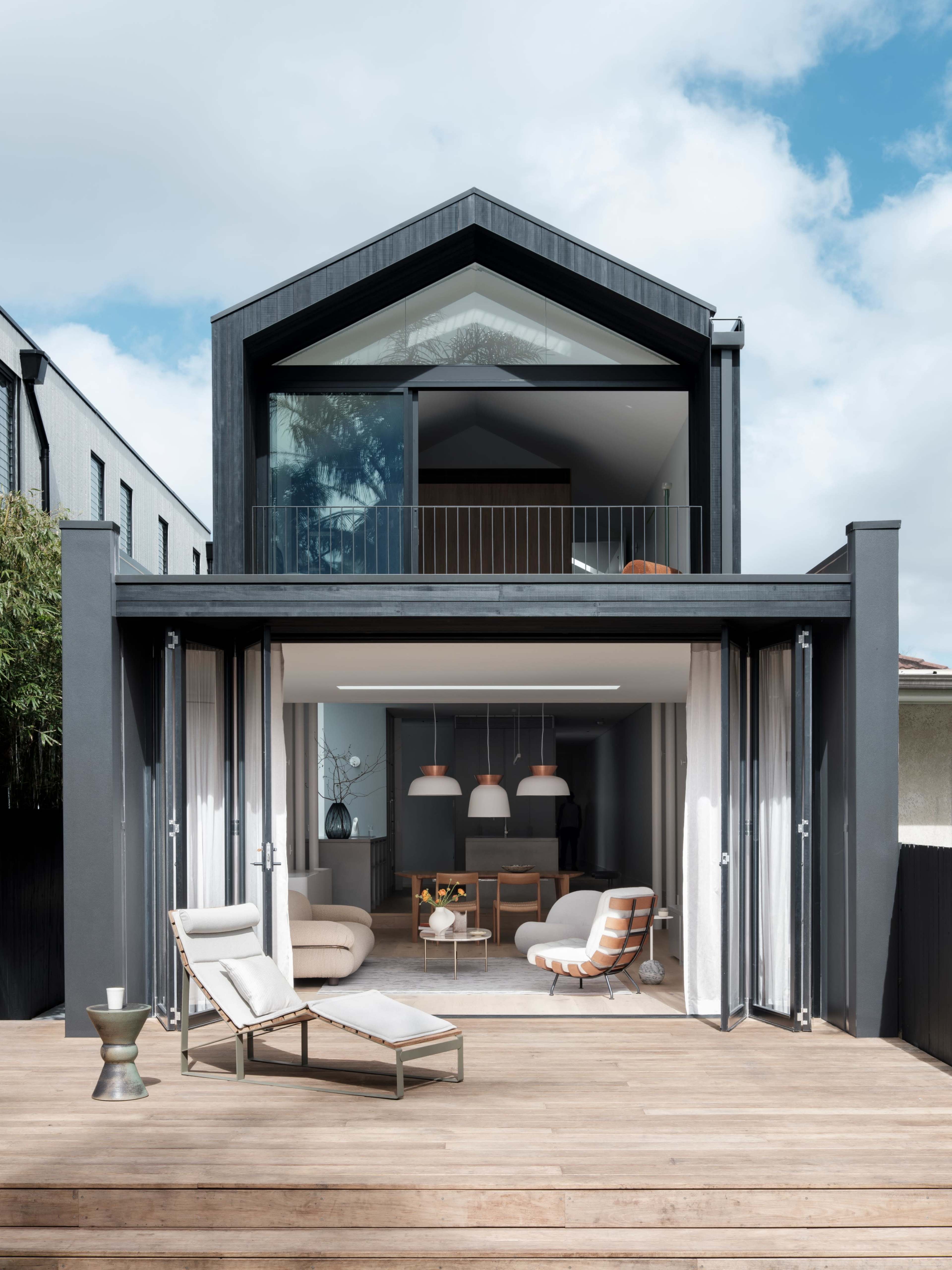 A modern two-story house with large glass doors opening onto a wooden deck, featuring minimalist furniture and pendant lighting inside.