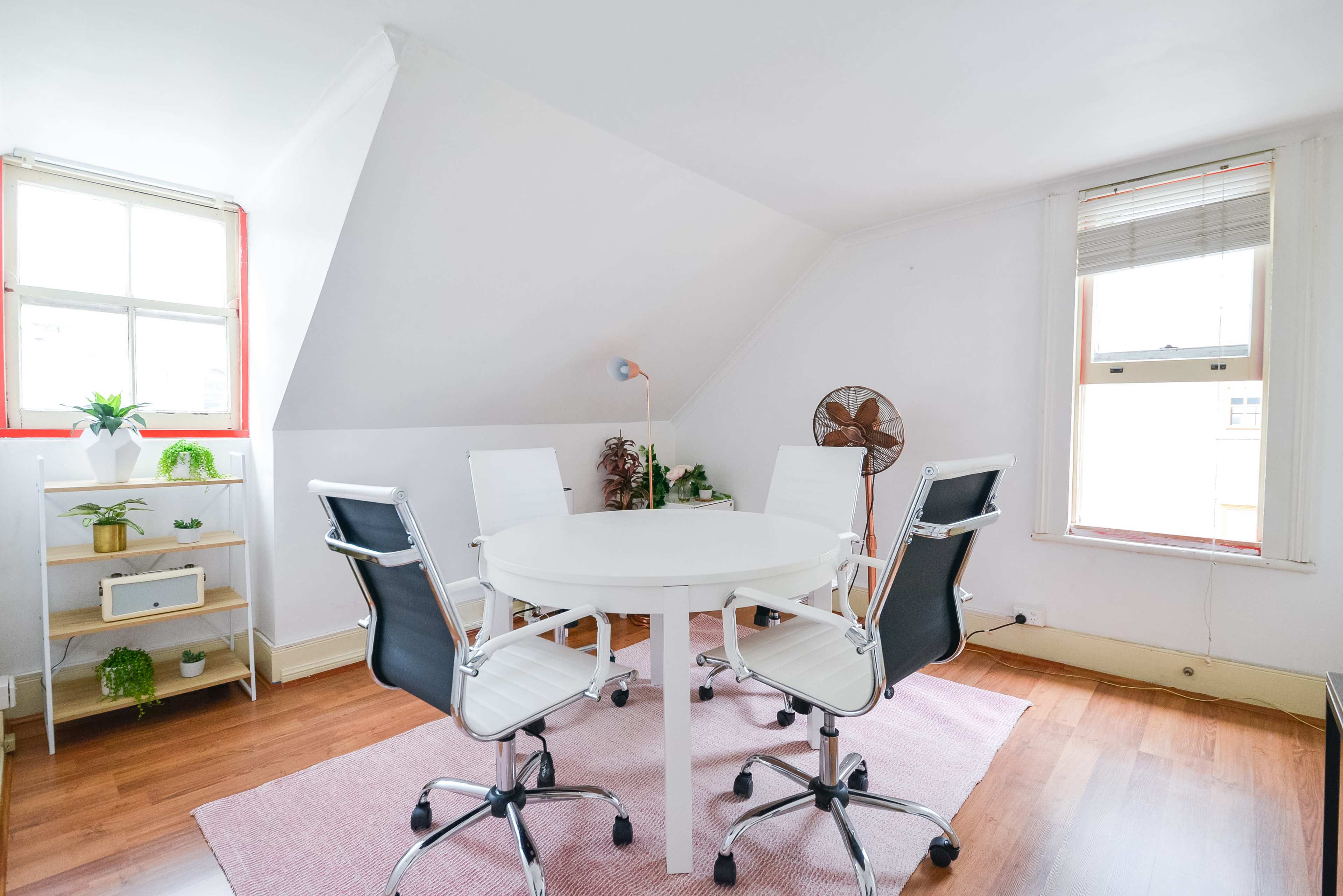 A small, bright room with a white round table surrounded by four black and white chairs, featuring a fan and a plant shelf against the walls.