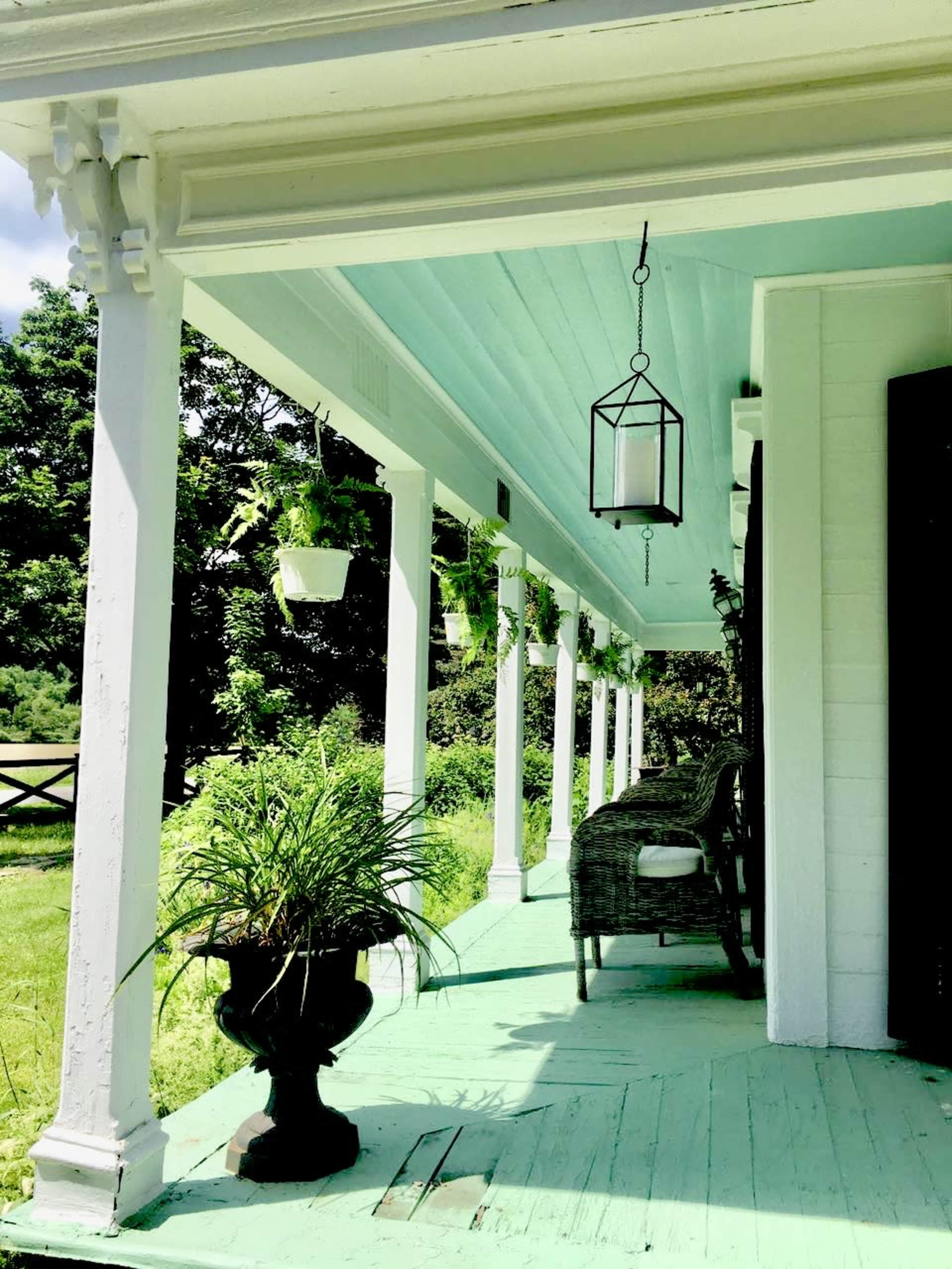 A porch with green-painted wood flooring, white columns, hanging planters, and a wicker chair beside a large black planter with greenery.