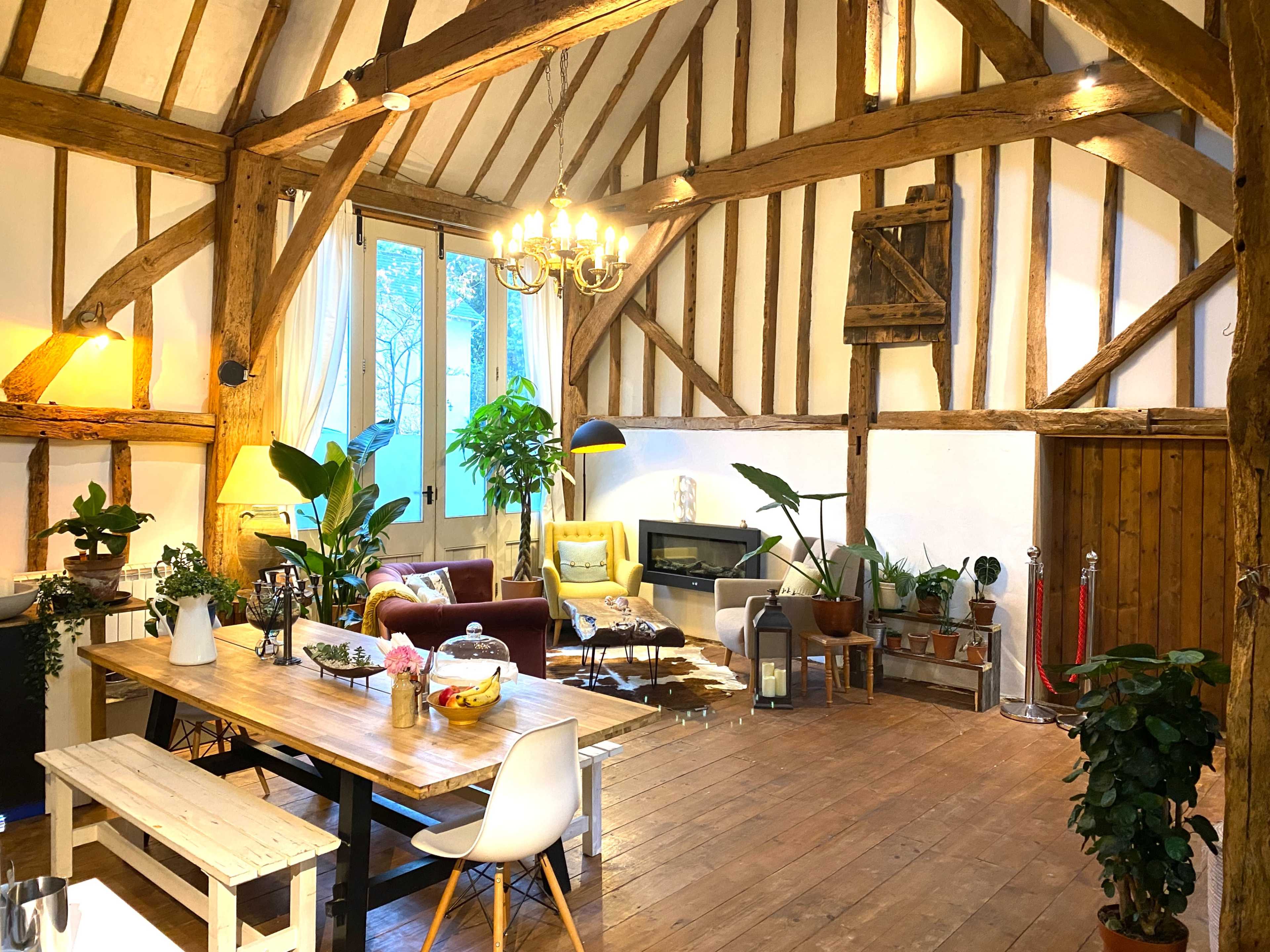 A spacious living area with wooden beams, a large dining table, various plants, and a cozy seating arrangement that includes a fireplace.