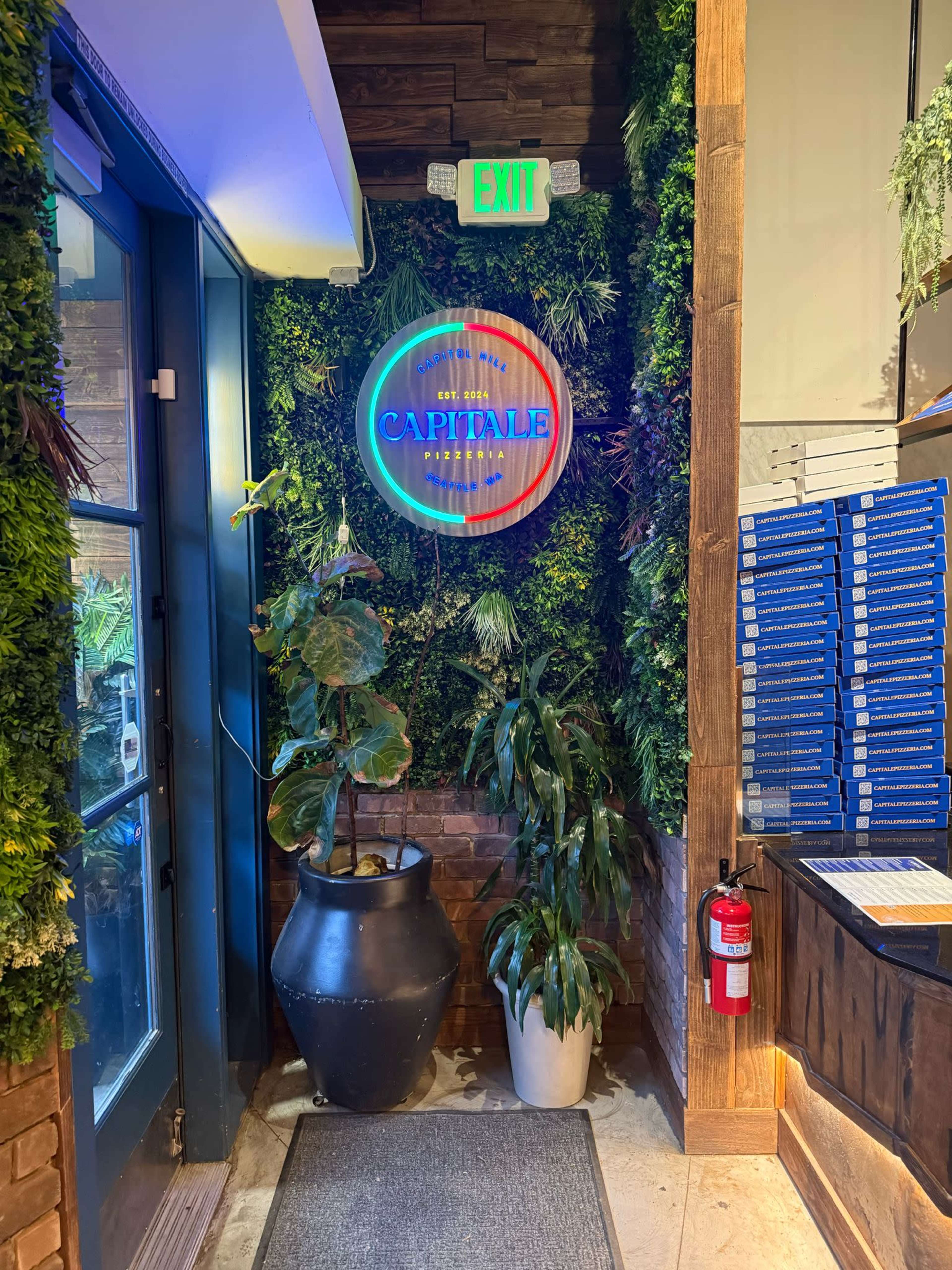 The image shows an indoor entrance with a neon sign reading "CAPITALE," surrounded by greenery and a large potted plant, leading to a menu display on the right.