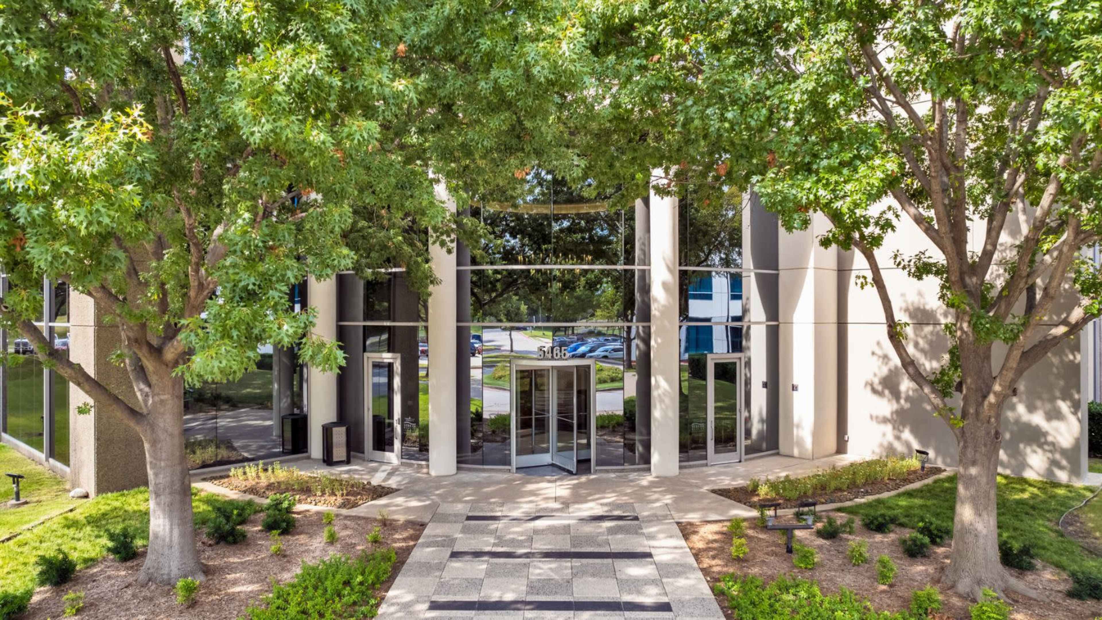 The entrance of a modern building features large glass doors flanked by two trees and a neatly landscaped pathway.