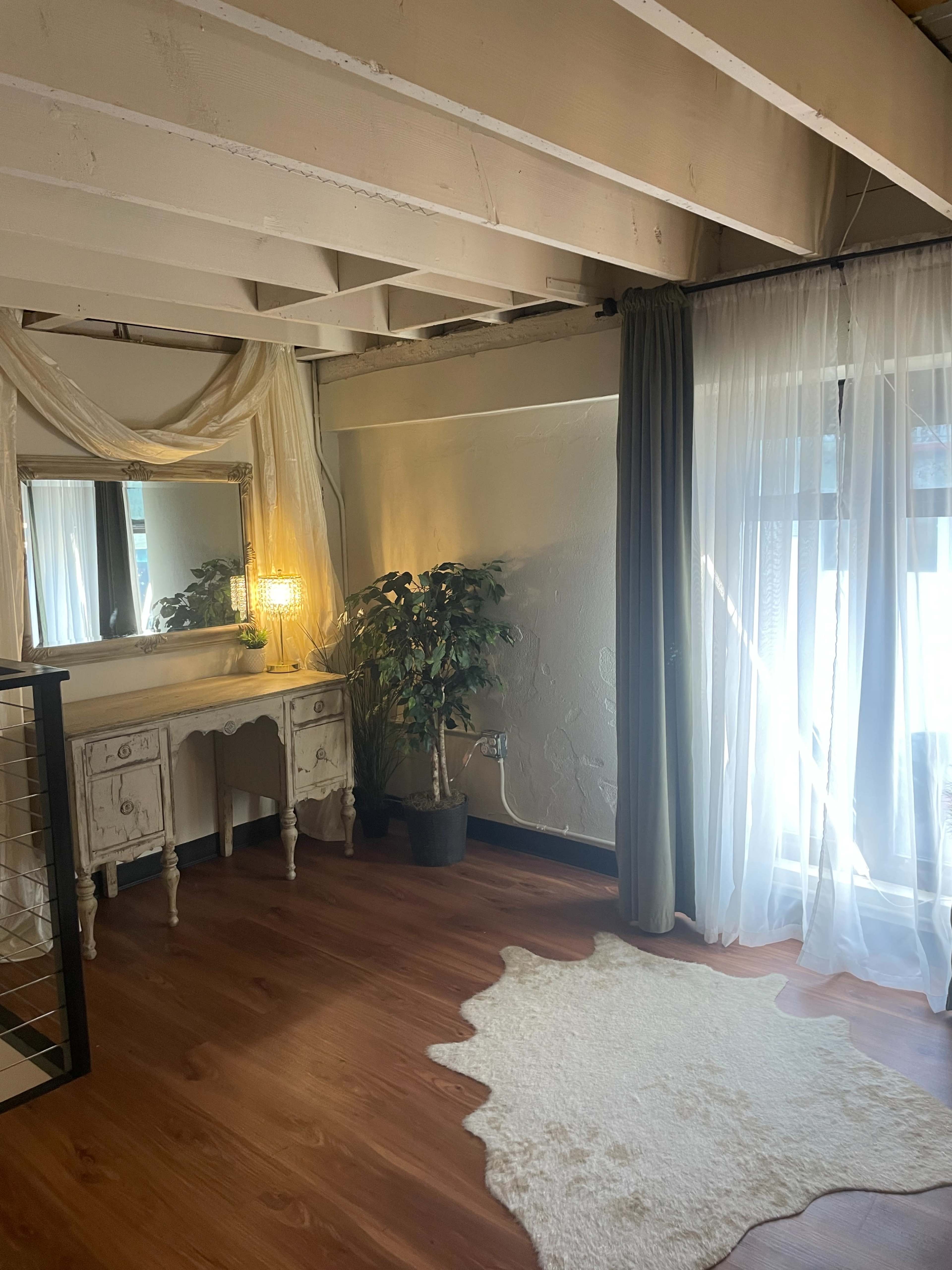 The image shows a well-lit interior space featuring a dressing table with a mirror, a decorative plant, and sheer curtains covering large windows.