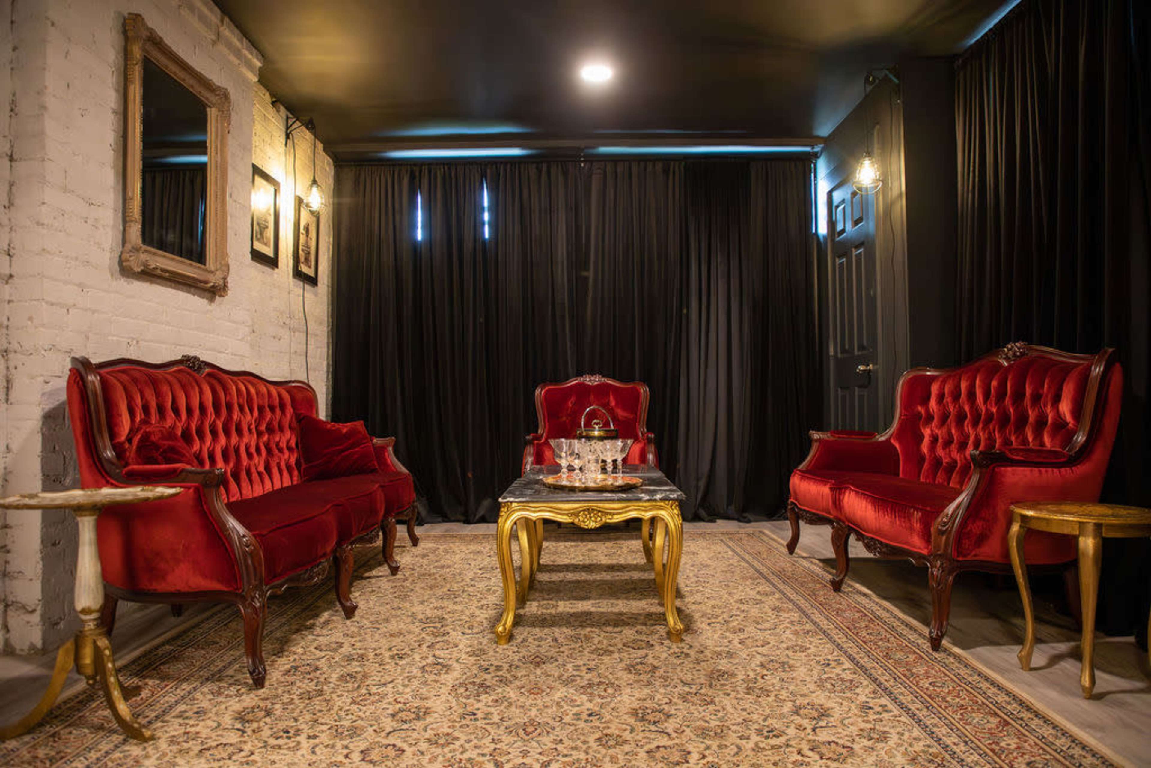 The image shows a sitting area with two red velvet sofas and a gold-accented coffee table on an ornate rug, surrounded by black curtains and framed mirrors on the walls.