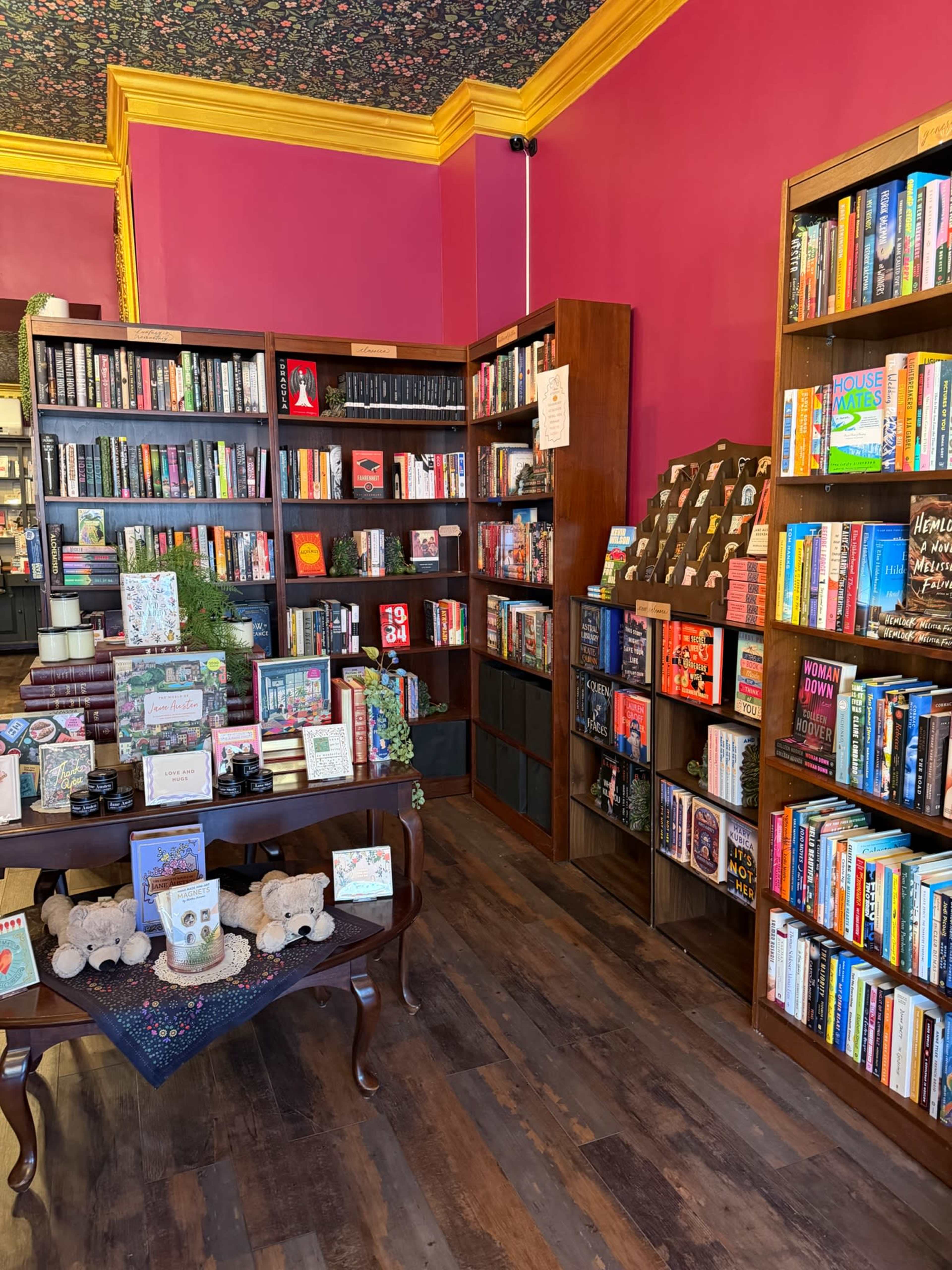 The image shows a cozy bookstore with two wooden bookshelves filled with various books, a decorative table displaying items, and vibrant pink walls.