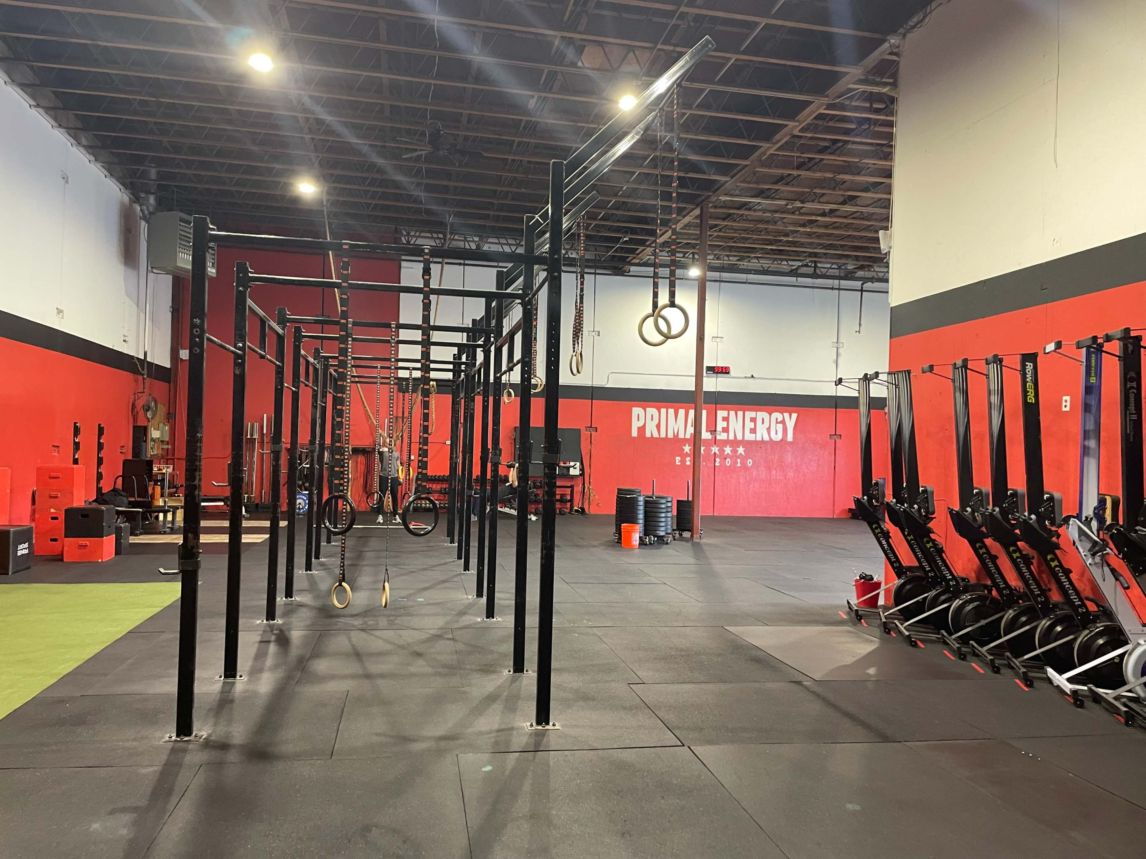 A gym interior with various fitness equipment, including pull-up bars, gymnastic rings, and rowing machines, against a backdrop of red and black walls.
