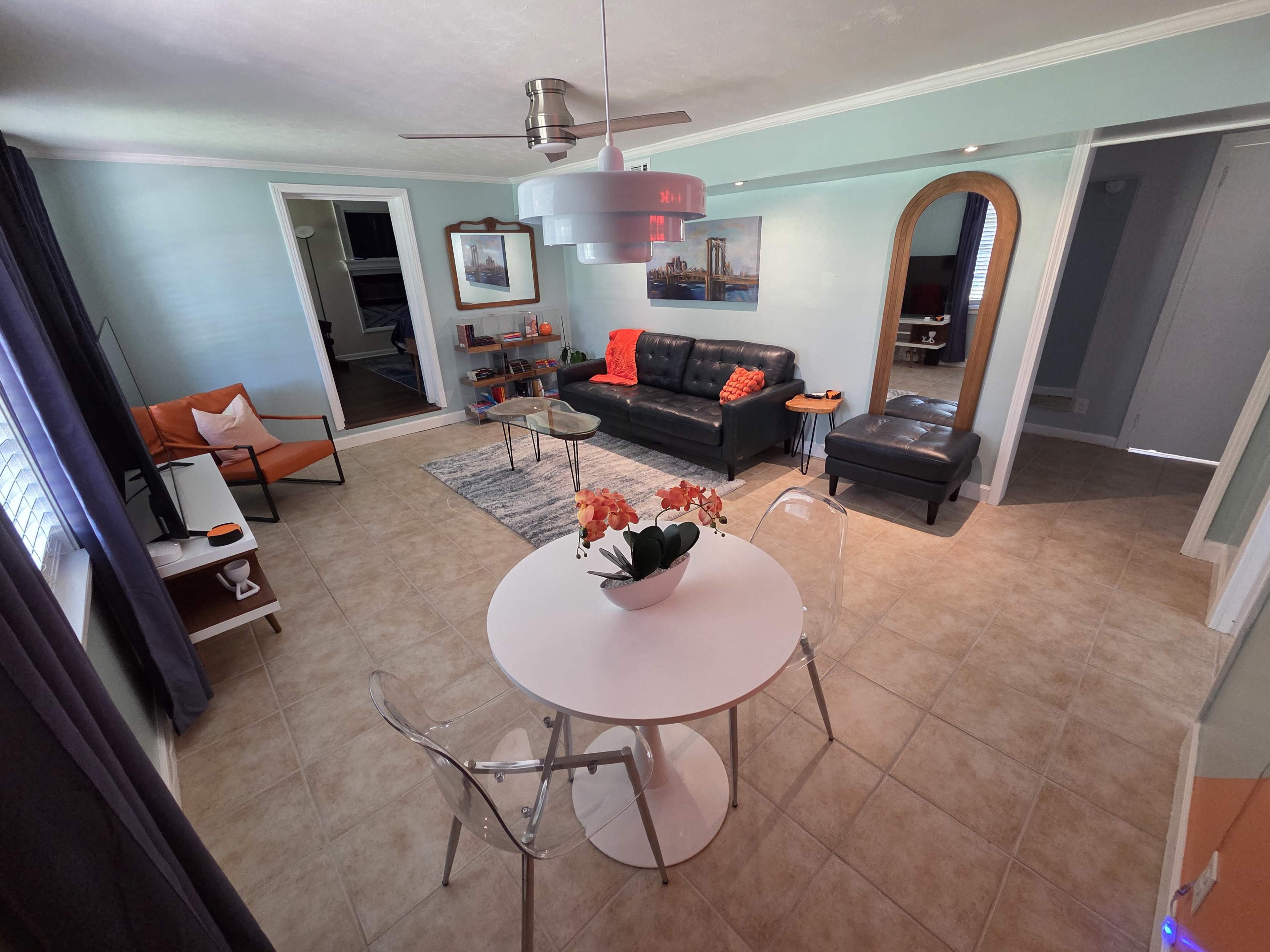 The brightly lit living room features a black couch, a white dining table with clear chairs, and a modern ceiling fan, with decorative elements like an orange throw and floral centerpiece.