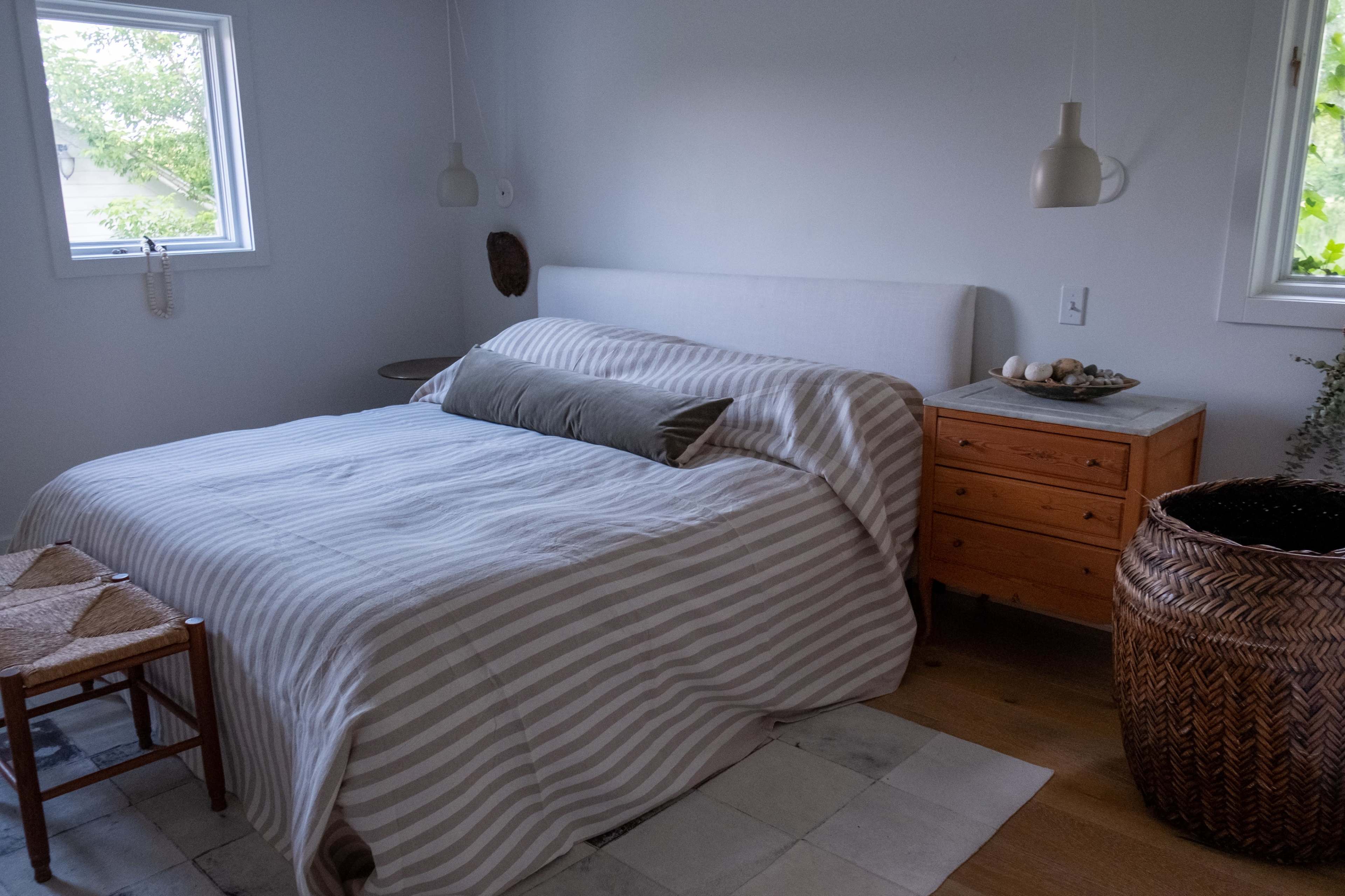 A minimalistic bedroom featuring a striped bedspread, a wooden nightstand with decorative items, and natural light streaming in through two windows.
