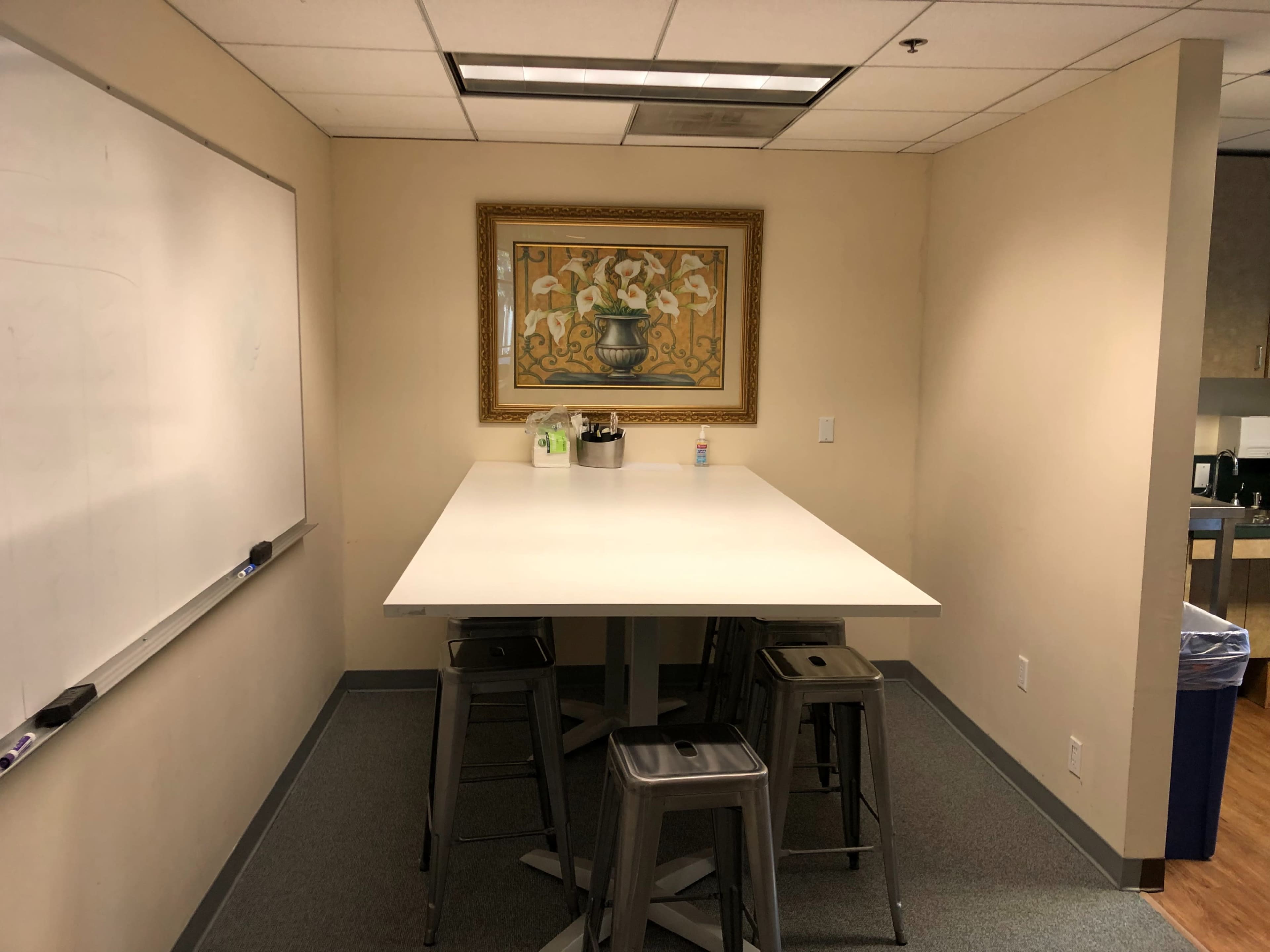 A small meeting area with a white table and metal bar stools, framed artwork on the wall, and a kitchenette in the background.