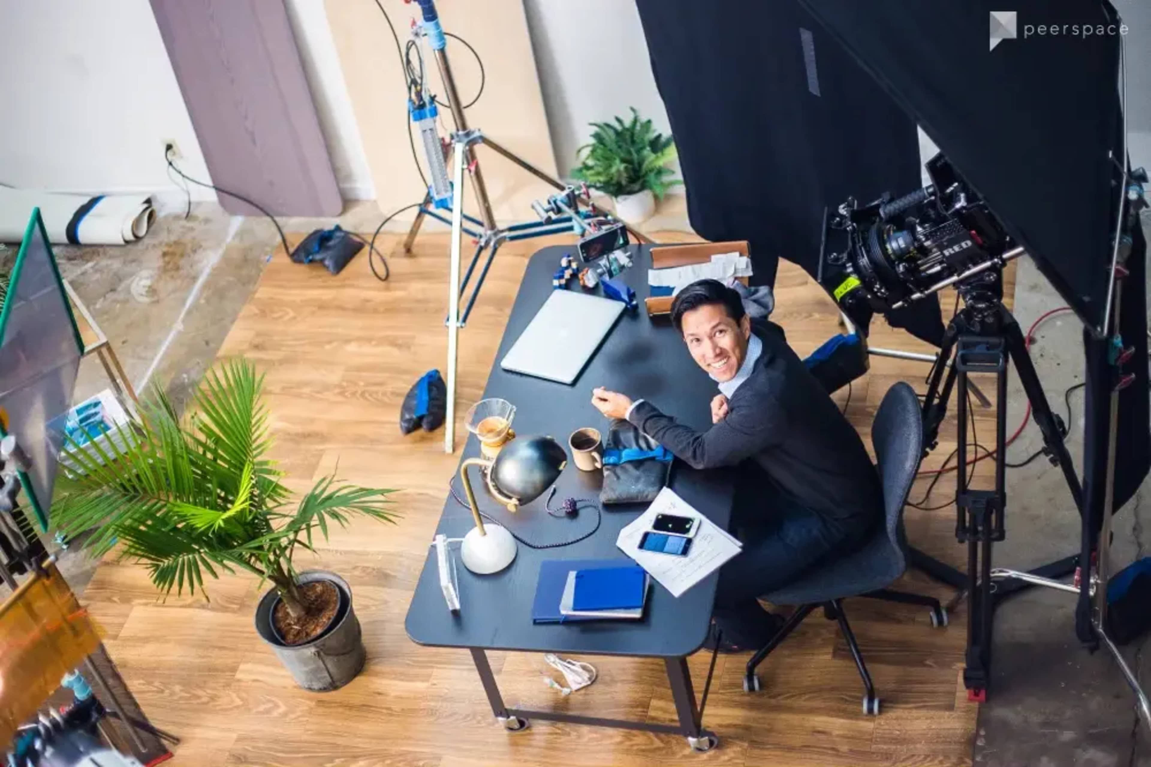 A person sits at a desk in a studio setup with a camera and lighting equipment nearby.