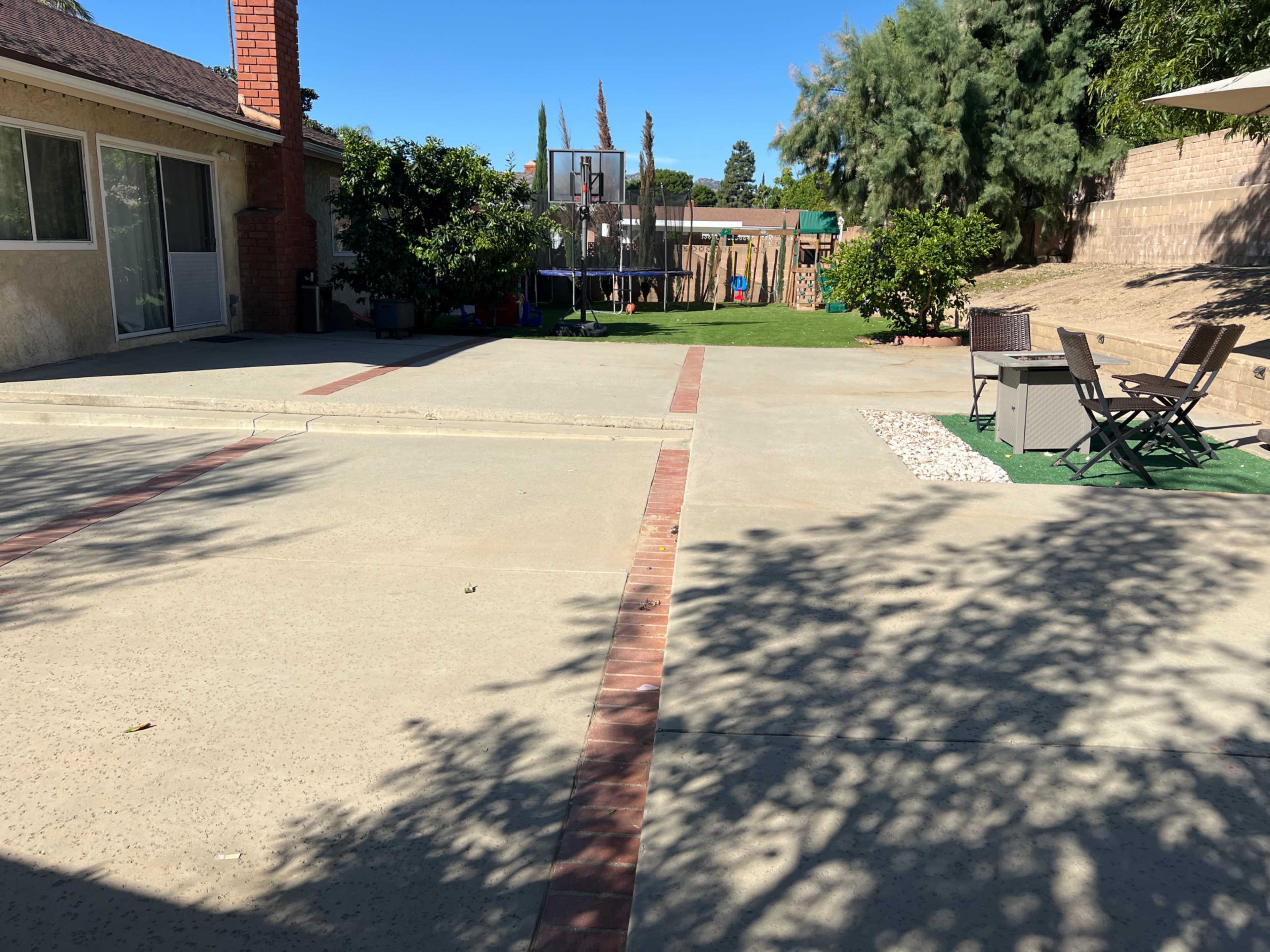 A spacious backyard with a concrete area, a basketball hoop, green grass, and seating arrangements under a shaded structure.