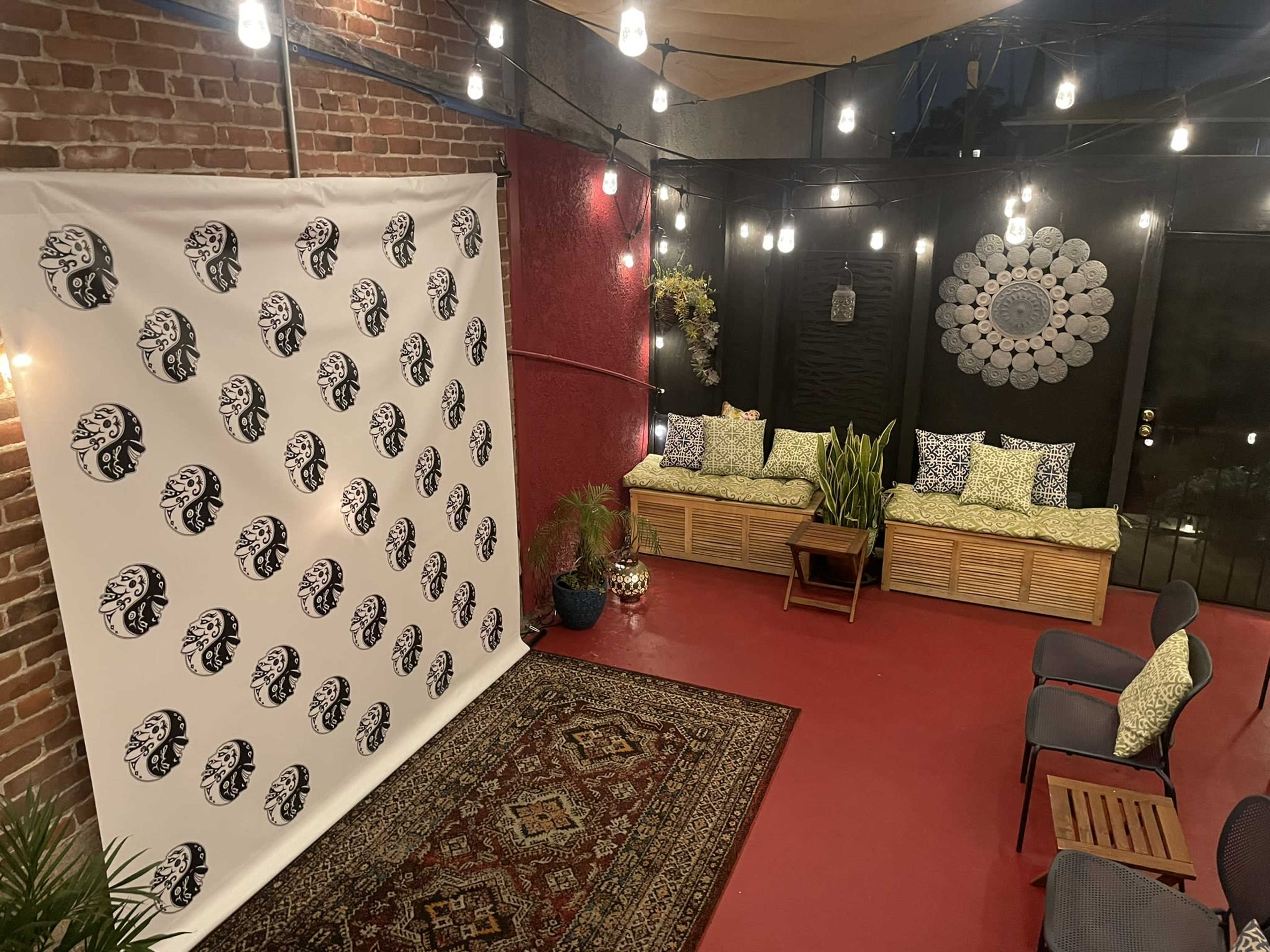 The image shows a cozy outdoor patio adorned with a patterned backdrop, wicker seating, plants, and string lights, set against a brick wall.