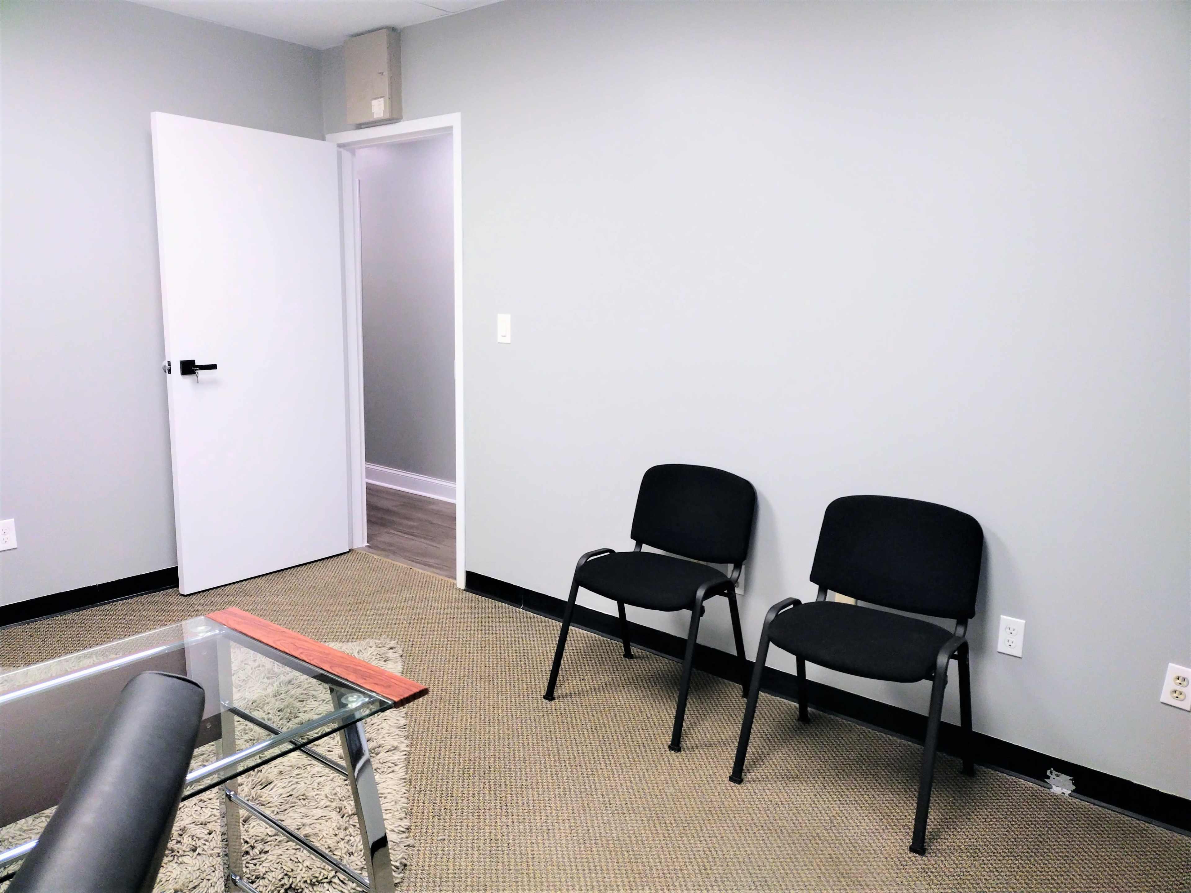 A small, empty room with two black chairs facing a glass table and a doorway leading to another area.