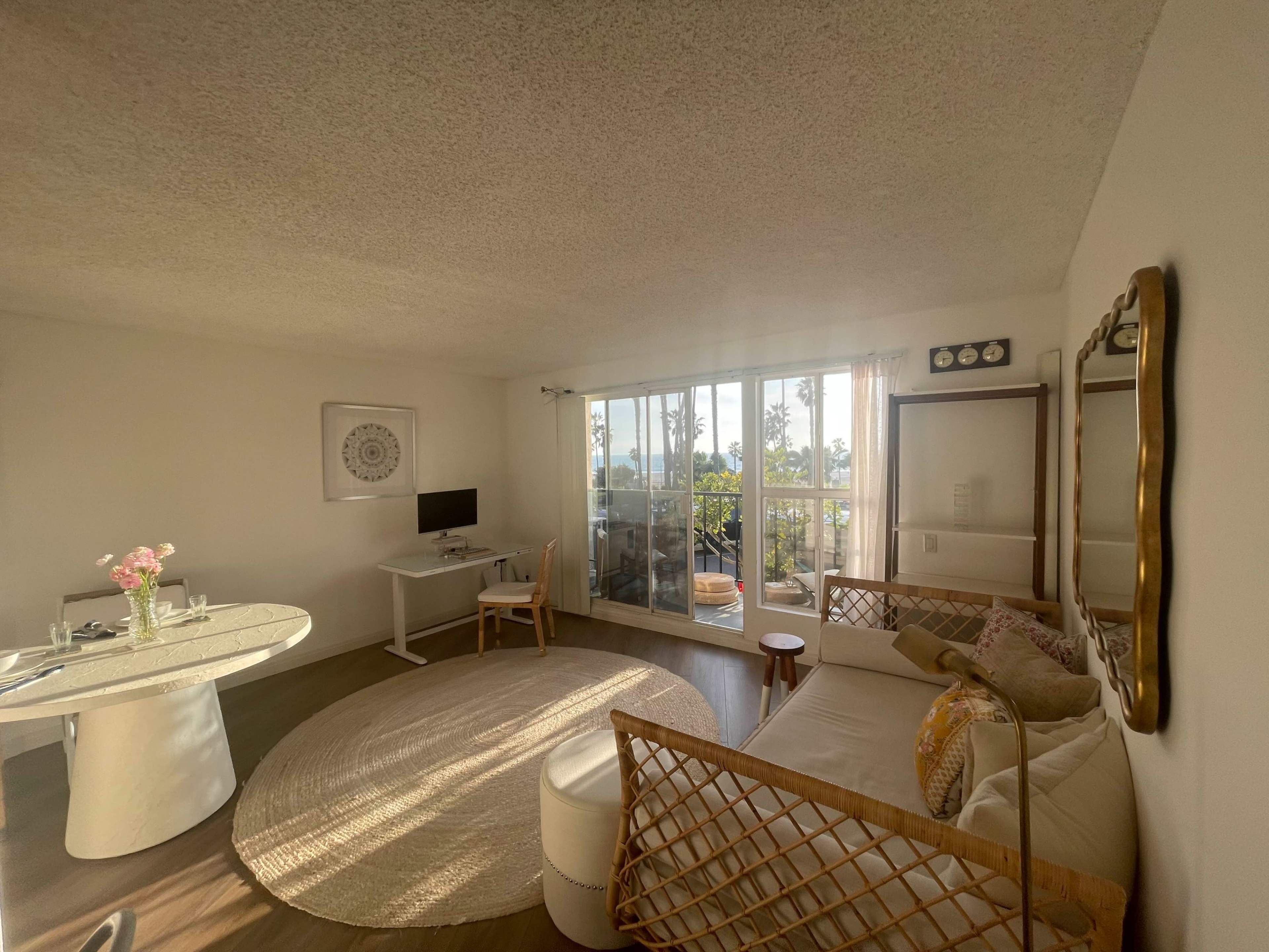 The image shows a bright living area with a circular dining table, a small work desk, a sofa, and large windows overlooking a balcony.