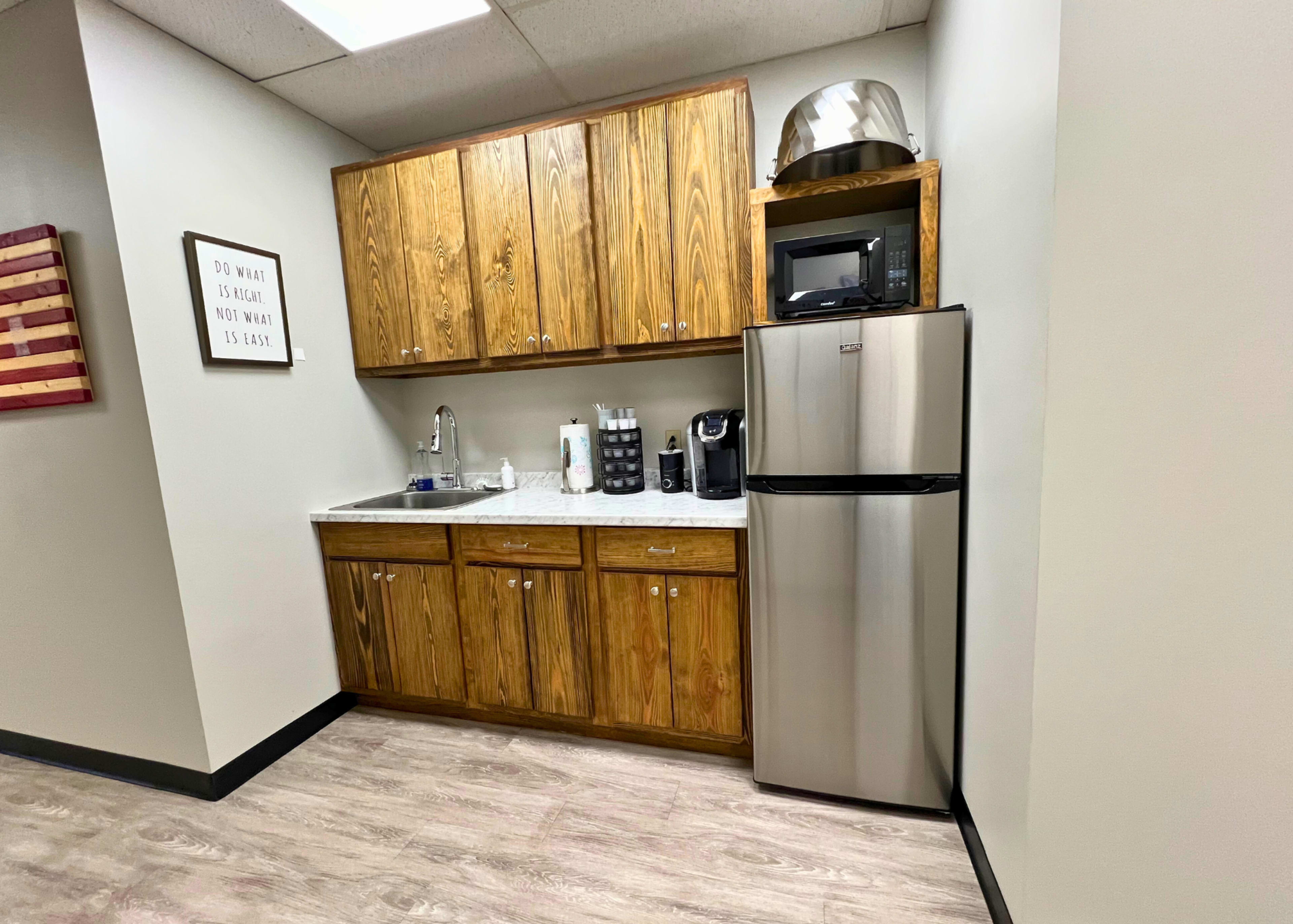 A small kitchen area with wooden cabinets, a countertop, a microwave, a coffee maker, and a stainless steel refrigerator.