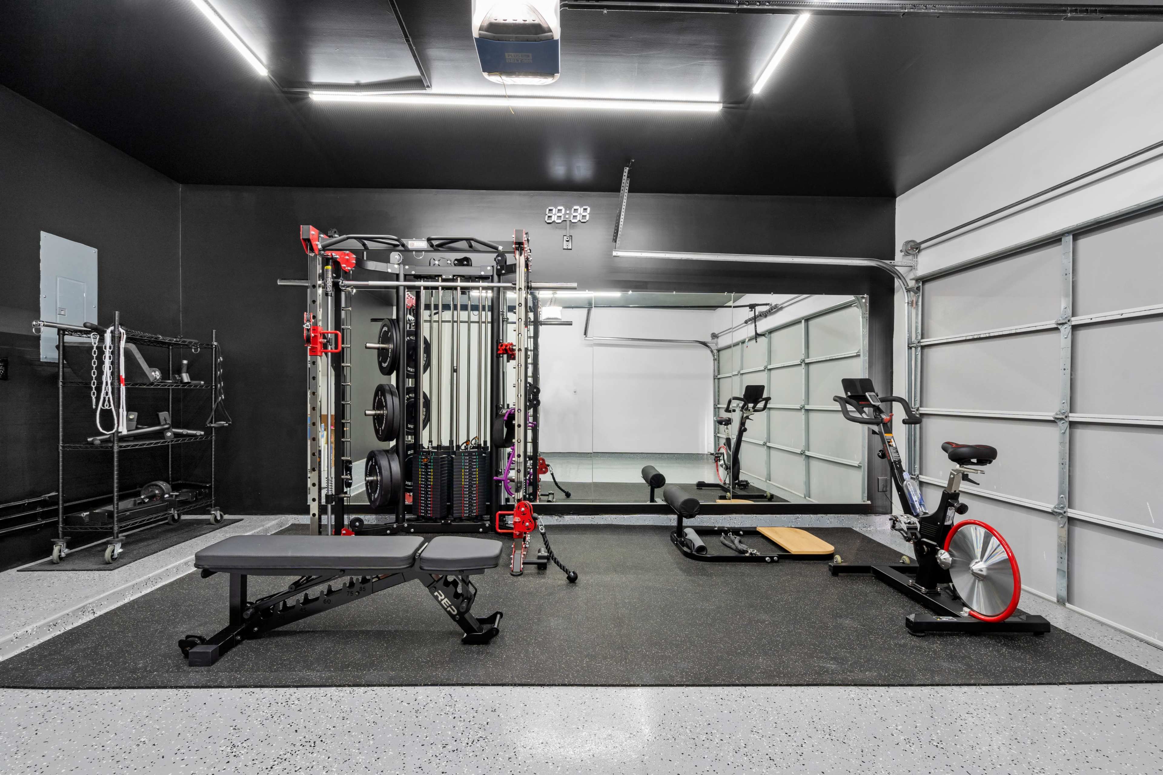 A modern home gym setup featuring a weight training station, a bench, a stationary bike, and exercise equipment arranged on a rubber floor mat in a garage space.
