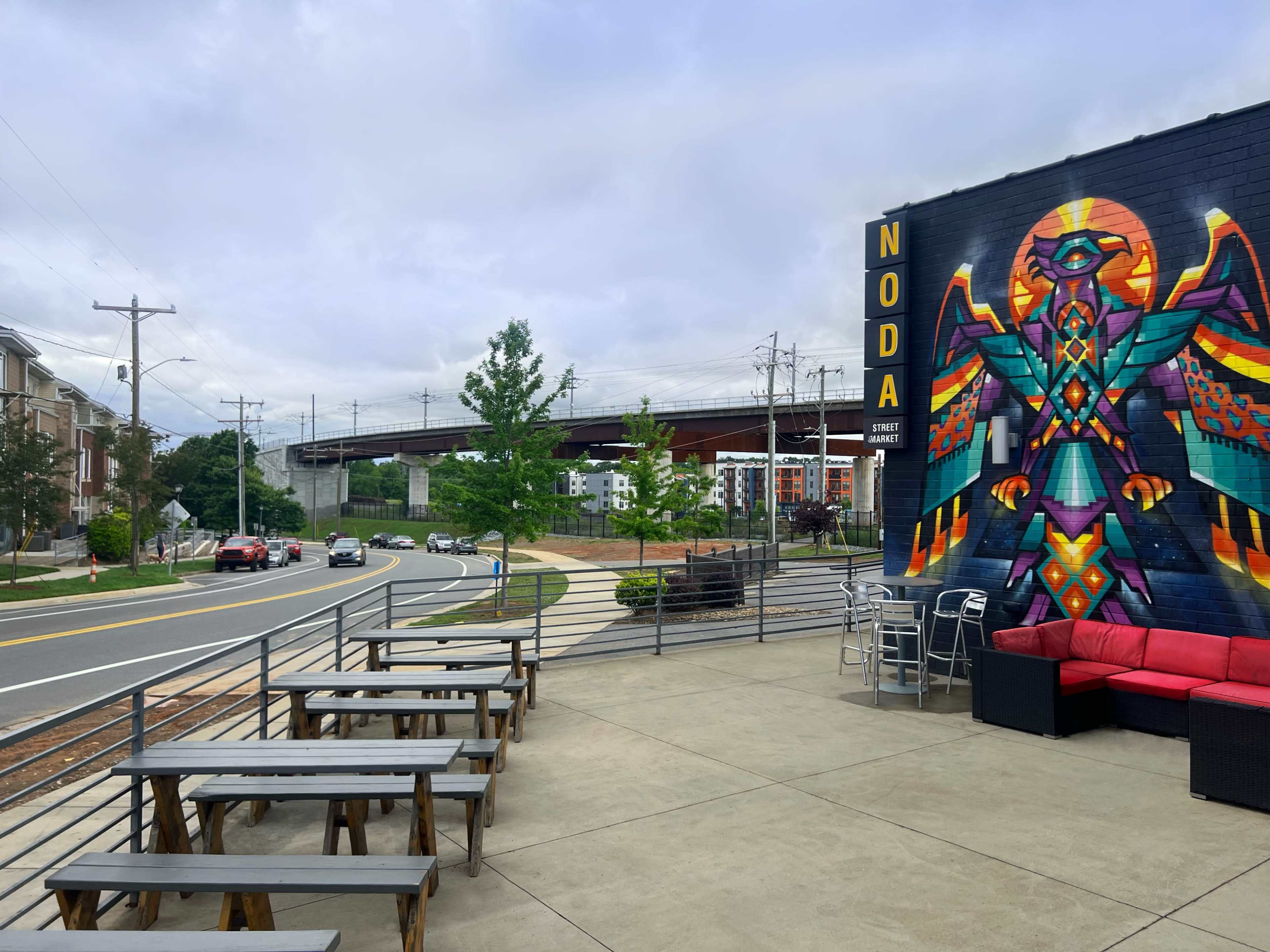An outdoor seating area with wooden tables in front of a mural featuring a colorful bird-like figure, next to a road and under an elevated train track.