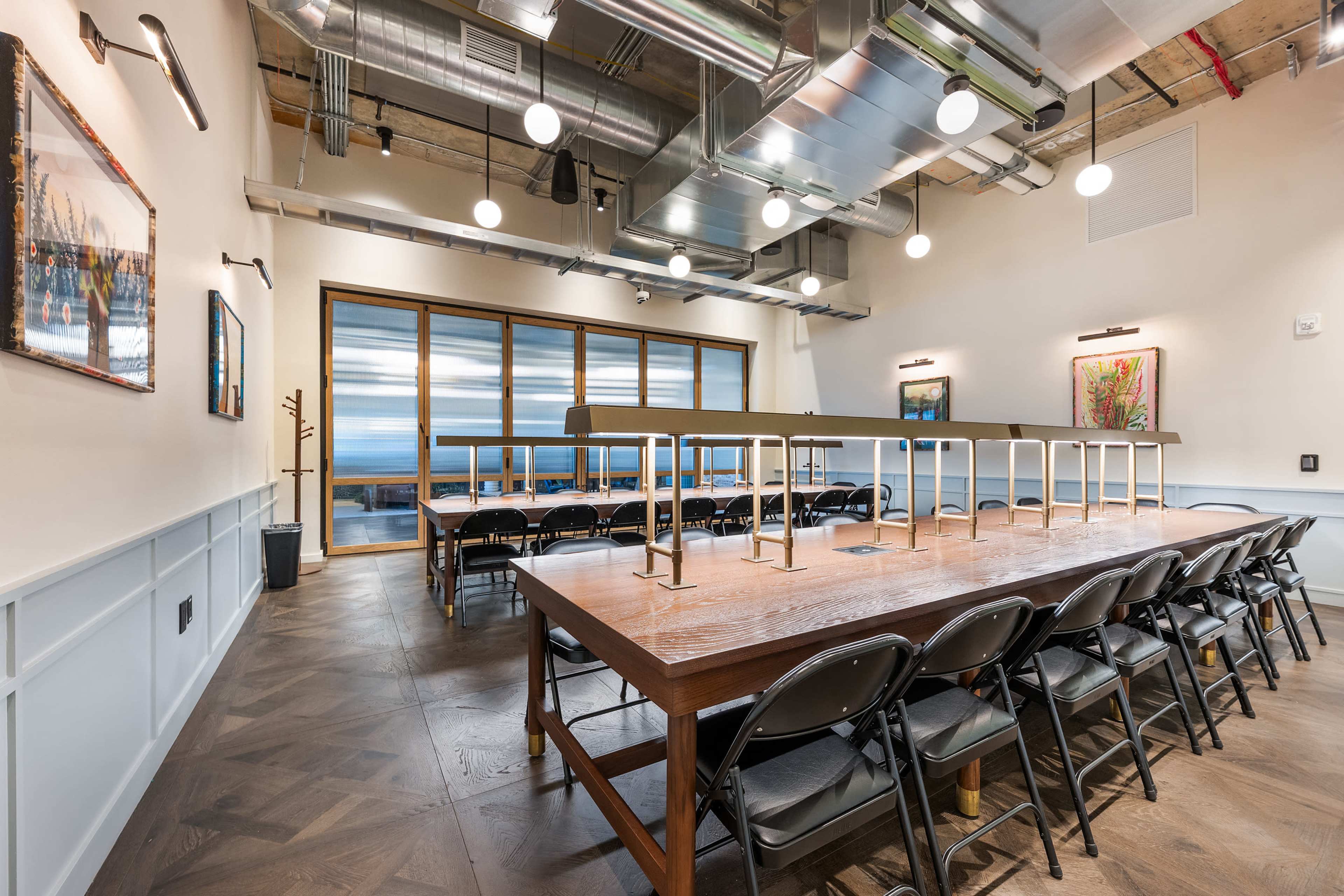 The image shows a well-lit conference room with a long wooden table surrounded by black folding chairs, large windows, and framed artworks on the walls.